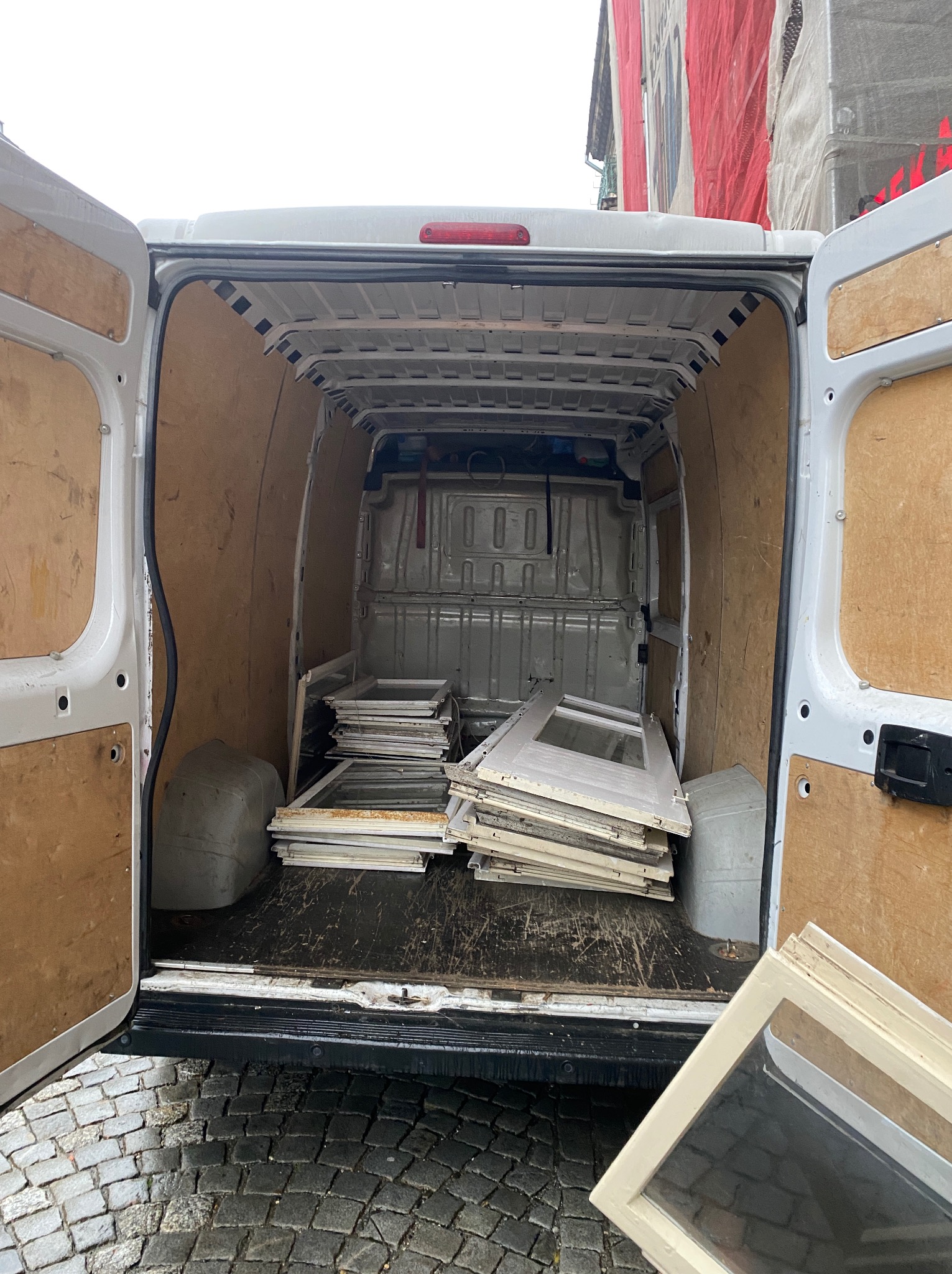 Otwarty furgon wypełniony starymi oknami, przygotowany do transportu, widok z tyłu na brukowaną ulicę. Czerwona siatka ochronna budynku w tle.