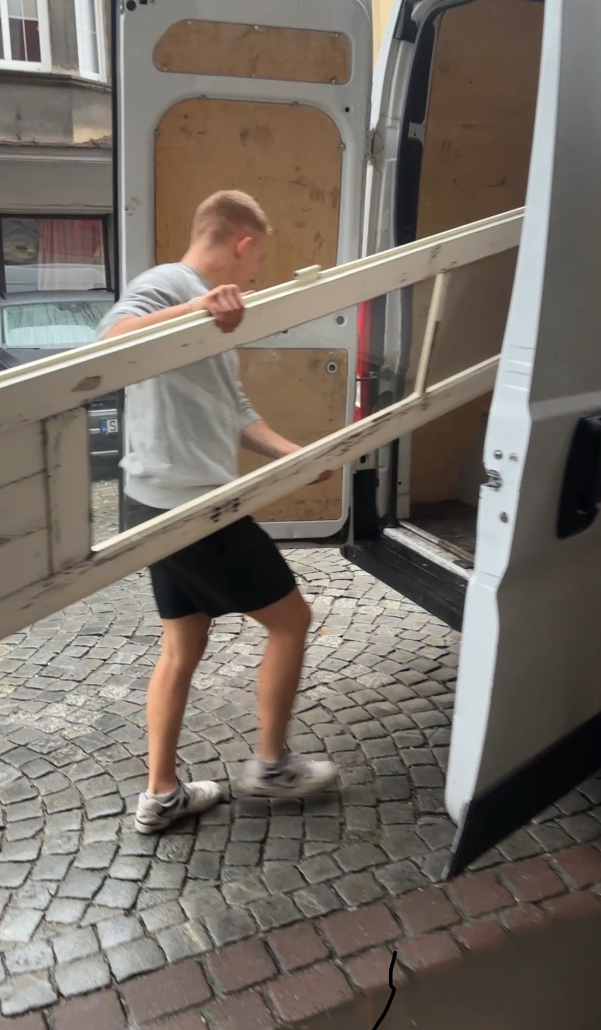 Mężczyzna wnosi białą, drewnianą ramę okienną do otwartego furgonetki na brukowanej ulicy. Widoczne drzwi furgonetki i fragment budynku w tle.
