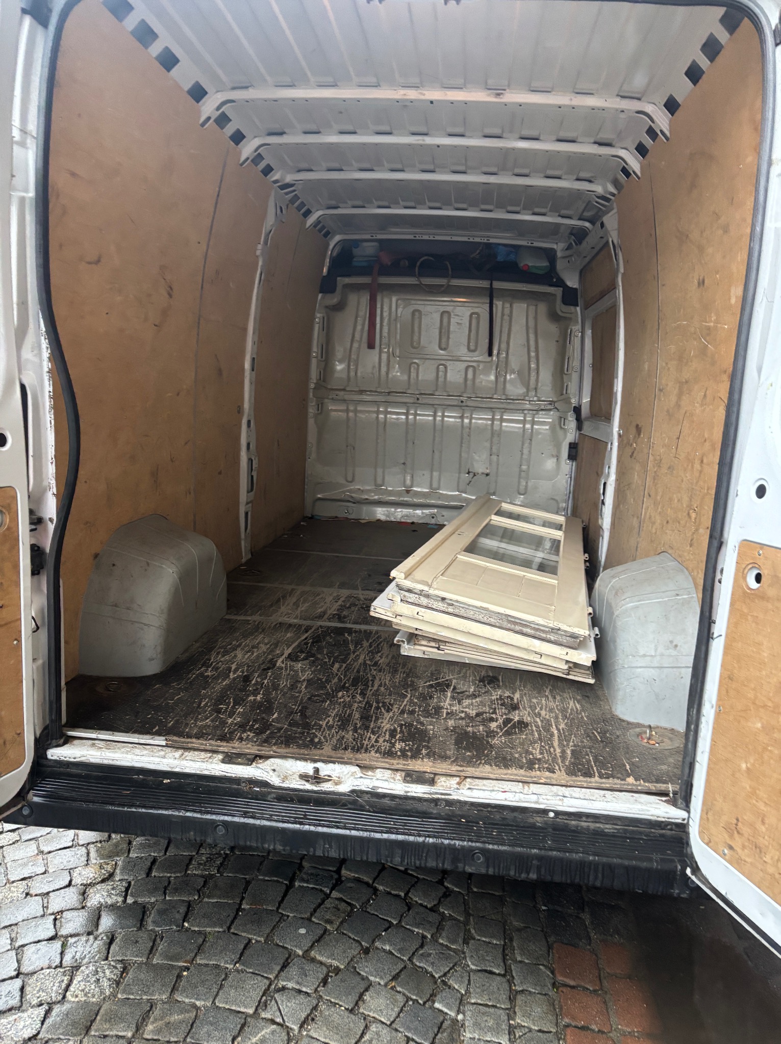Wnętrze furgonetki z drewnianą zabudową, przygotowanej do transportu drzwi. Widoczna podłoga z śladami użytkowania oraz fragment brukowanej ulicy.