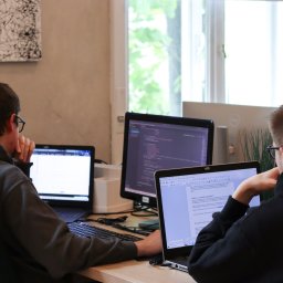 MinisterstwoReklamy.pl Sp. z o.o. - Dwóch programistów pracuje przy biurkach w biurze. Jeden pisze kod, drugi edytuje tekst. Na biurkach monitory i laptopy. W tle okno i abstrakcyjny obraz.