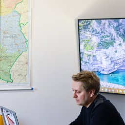 MinisterstwoReklamy.pl Sp. z o.o. - Mężczyzna z tatuażem na przedramieniu pracuje na laptopie. W tle mapa Polski i monitor z widokiem na malownicze miasto nad morzem. Laptop ozdobiony naklejkami.