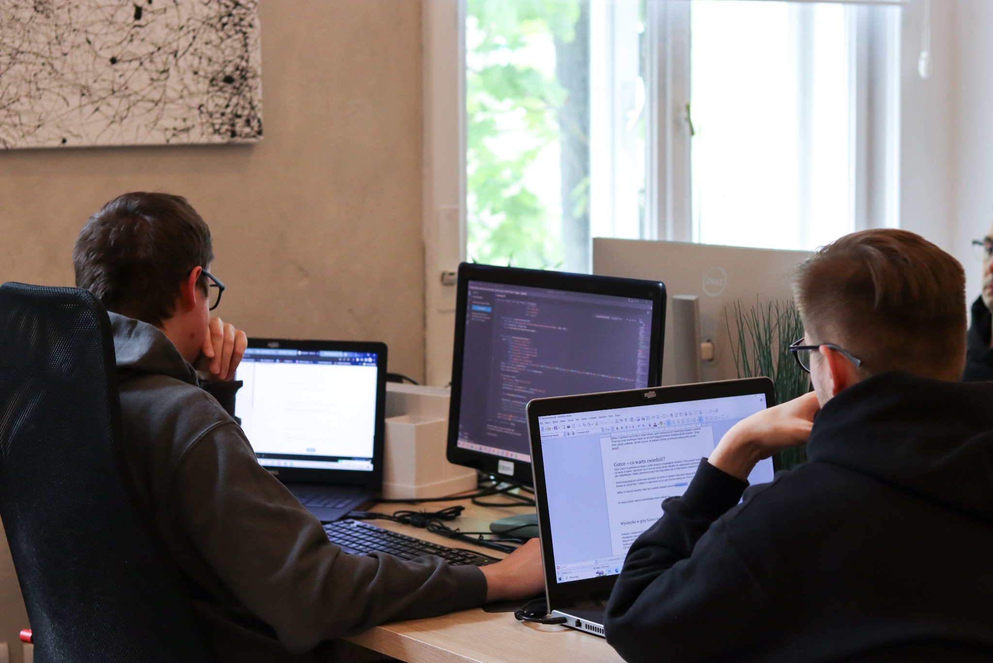 Dwóch programistów pracuje przy biurkach w biurze. Jeden pisze kod, drugi edytuje tekst. Na biurkach monitory i laptopy. W tle okno i abstrakcyjny obraz.
