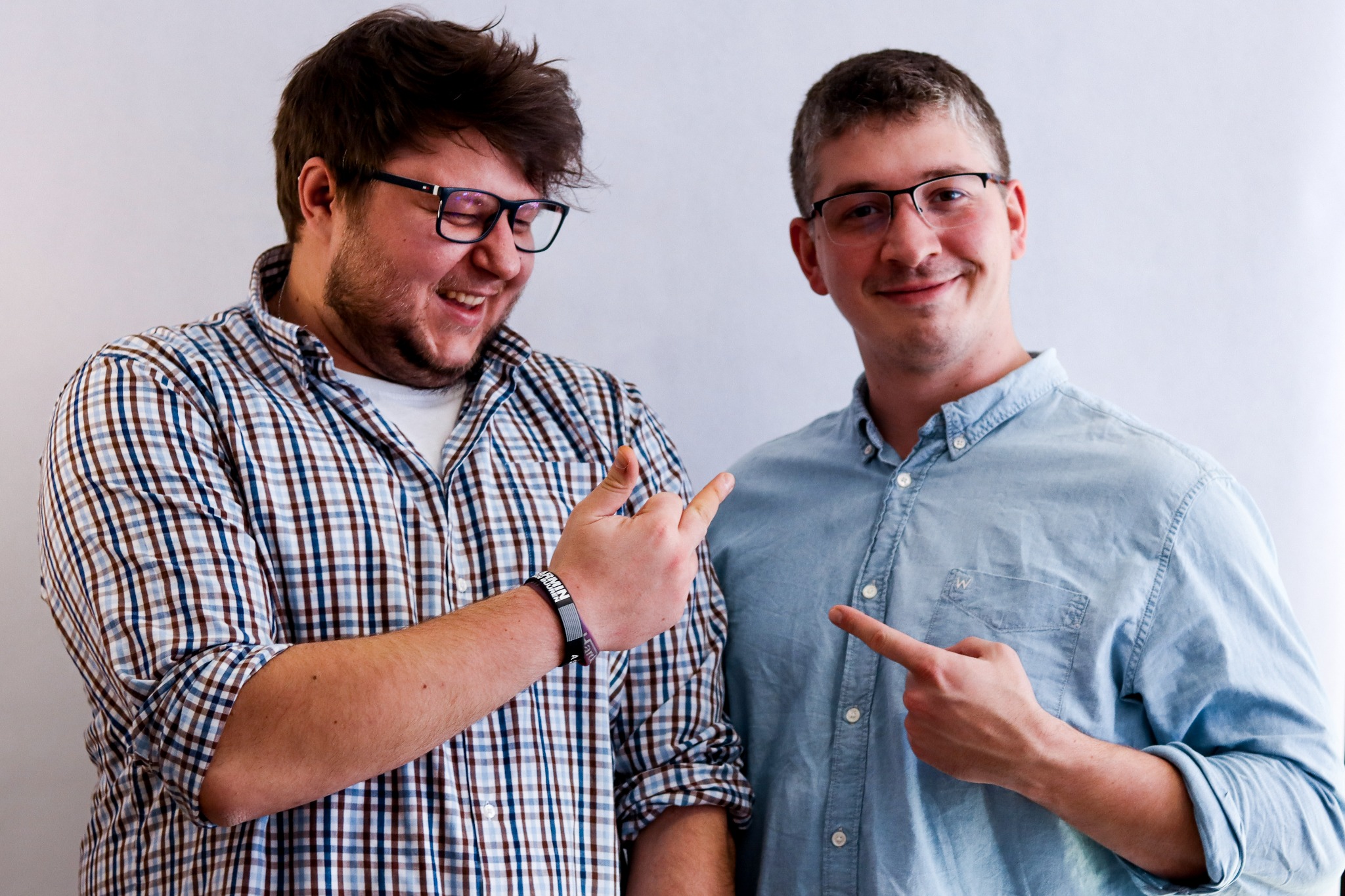 Dwóch uśmiechniętych mężczyzn w okularach wskazuje palcami w dół. Jeden w kraciastej koszuli, drugi w jeansowej. Jasne tło. Zespół, współpraca, prezentacja.