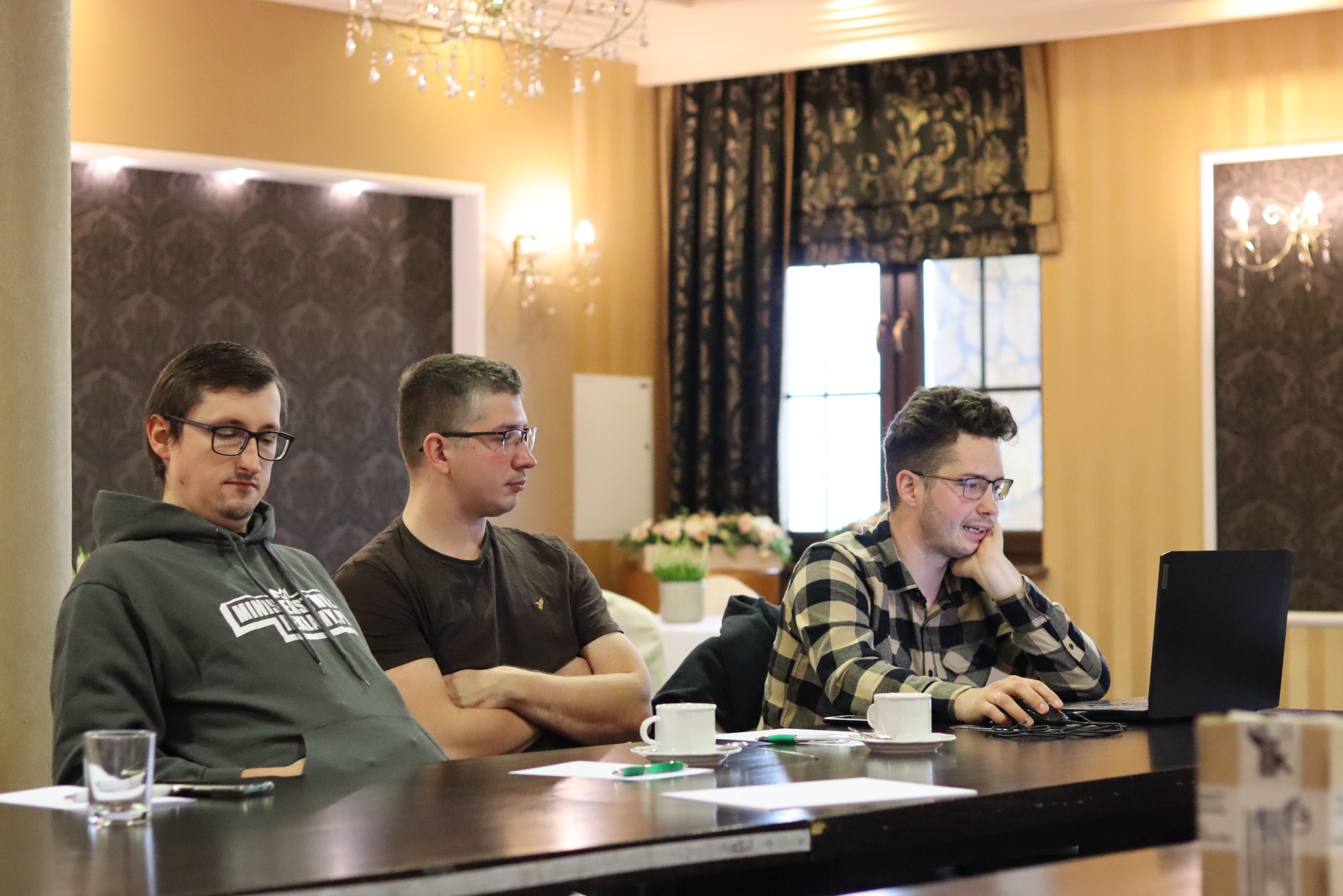 Trzech mężczyzn w okularach siedzi przy stole z laptopem i kawą w eleganckim wnętrzu. Jeden z nich pracuje na laptopie, dwaj pozostali obserwują.