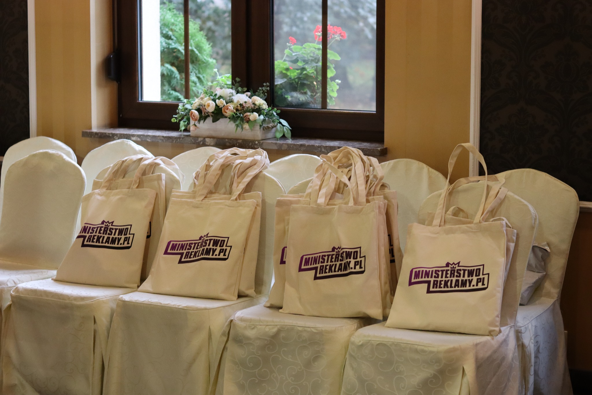 Beżowe torby bawełniane z logo 'Ministerstwo Reklamy.pl' ustawione na krzesłach z białymi pokrowcami, w tle okno z kwiatami.