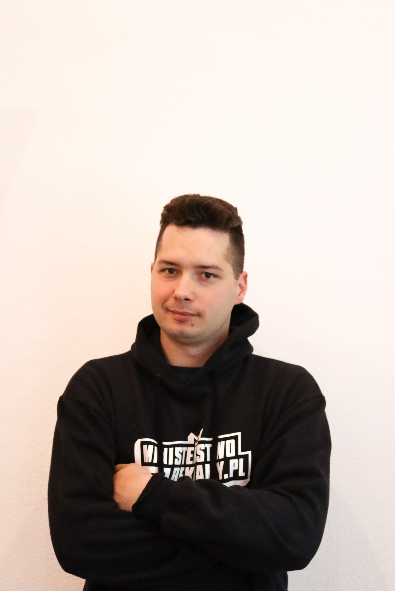 Młody mężczyzna w czarnej bluzie z nadrukiem, stoi na jasnym tle, krzyżując ręce. Uśmiechnięty, patrzy wprost. Widoczny od ramion w górę.