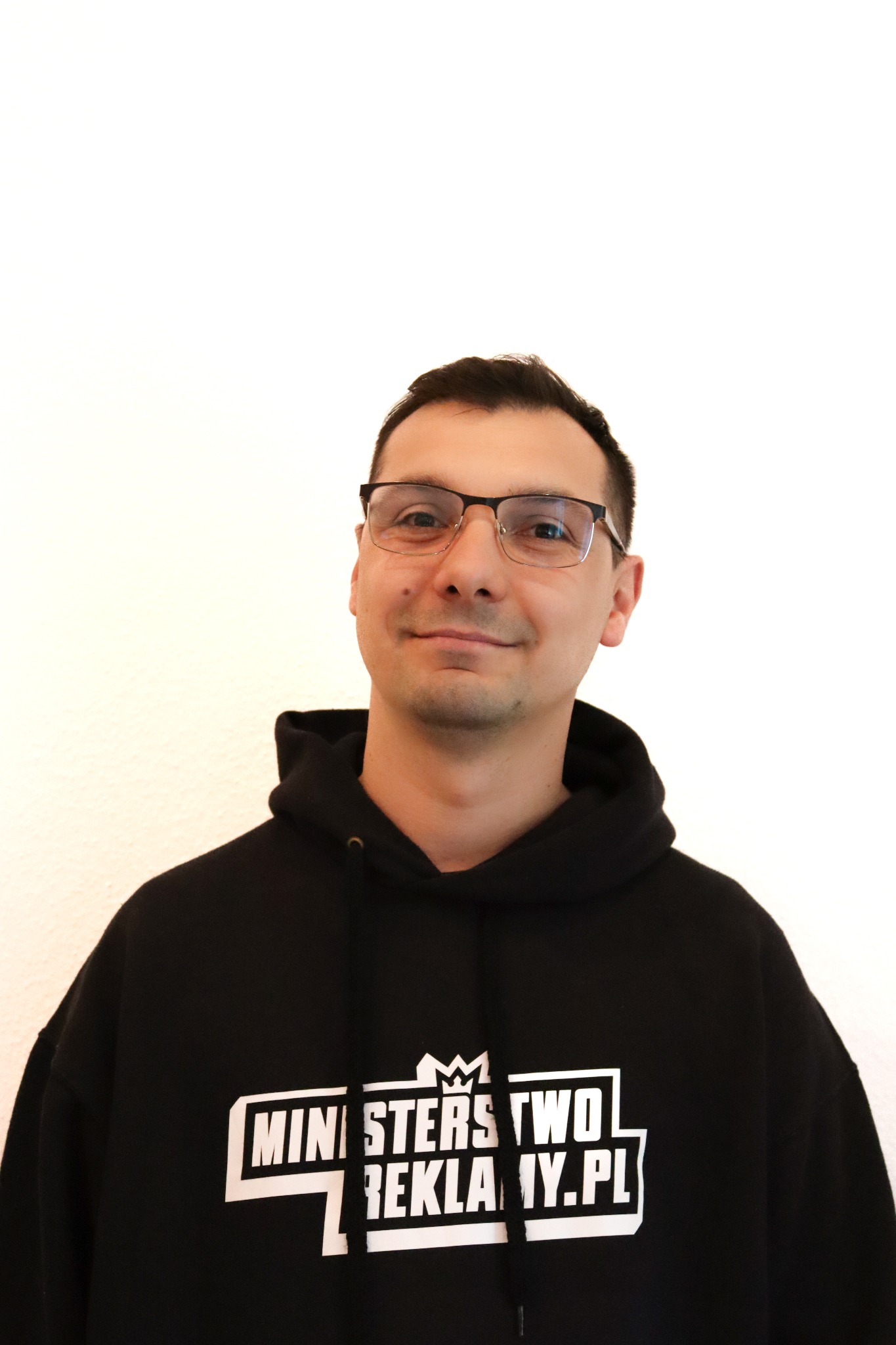 Mężczyzna w czarnej bluzie z kapturem i logo 'Ministerstwo Reklamy.pl' na białym tle. Uśmiechnięty, w okularach. Portret.