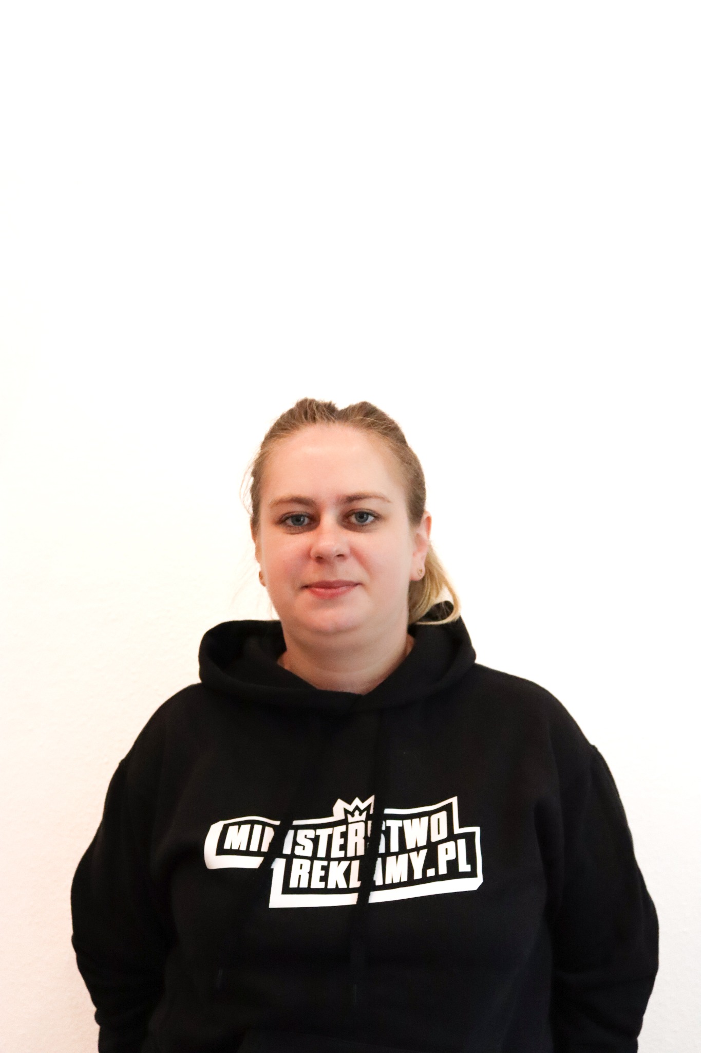 Portret kobiety w czarnej bluzie z logo 'Ministerstwo Reklamy.pl' na jasnym tle. Uśmiechnięta, z blond włosami upiętymi do tyłu. Ujęcie od pasa w górę.