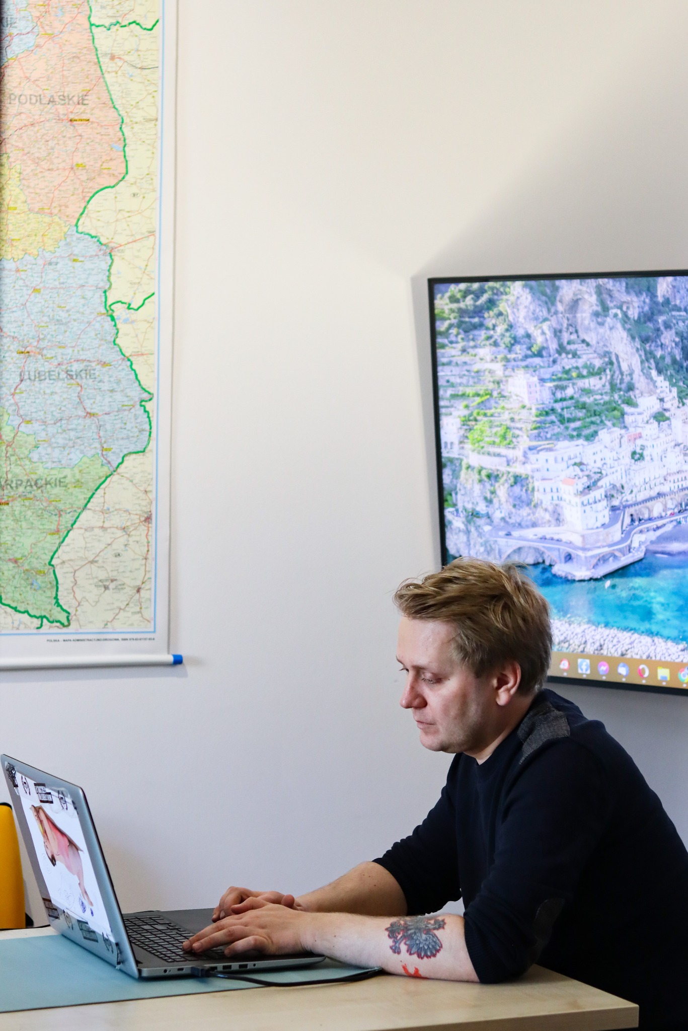 Mężczyzna z tatuażem na przedramieniu pracuje na laptopie. W tle mapa Polski i monitor z widokiem na malownicze miasto nad morzem. Laptop ozdobiony naklejkami.
