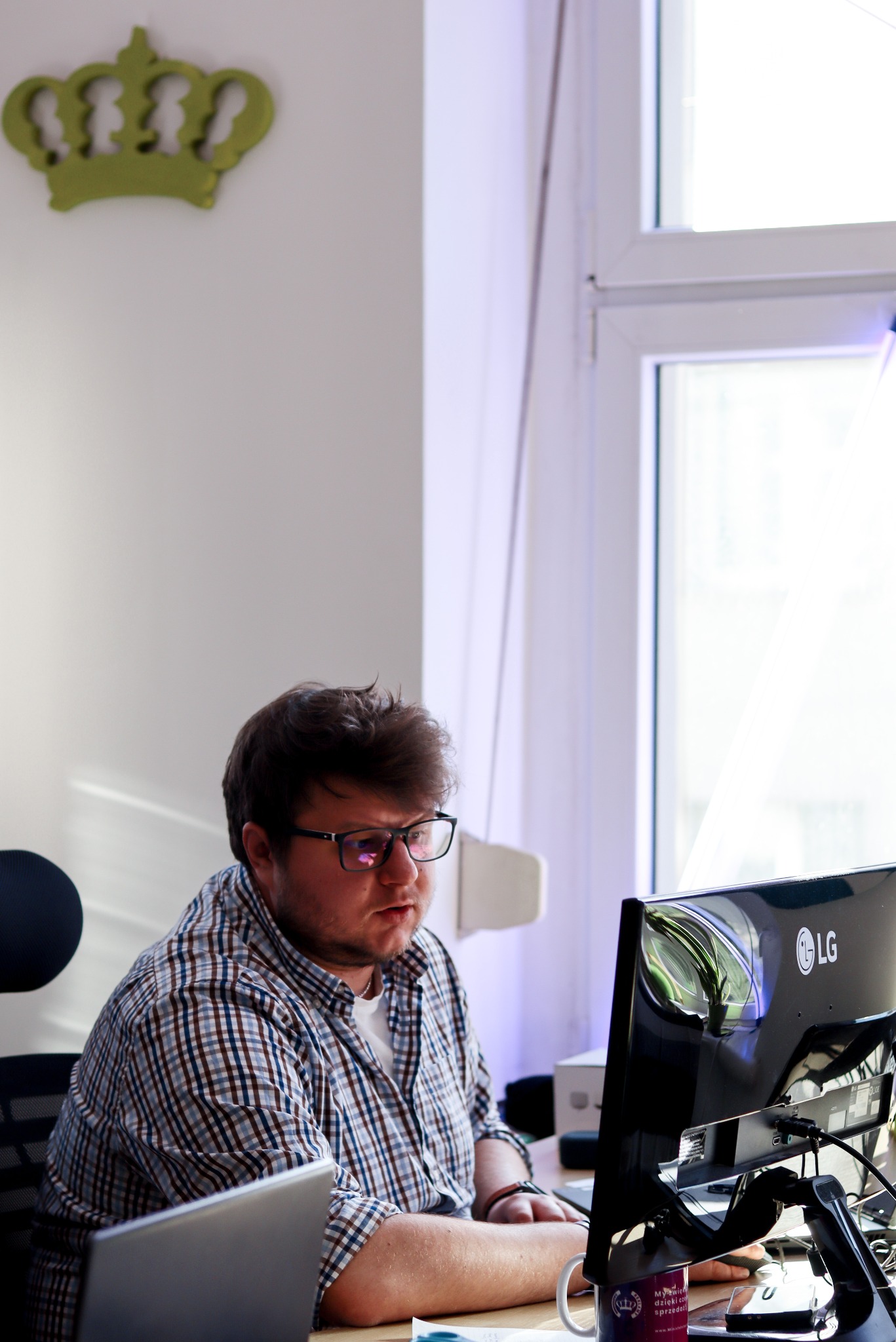 Mężczyzna w okularach pracuje przy biurku z monitorem LG. Na ścianie zielona korona, na biurku kubek z logo. Widoczne laptop i inne akcesoria biurowe.