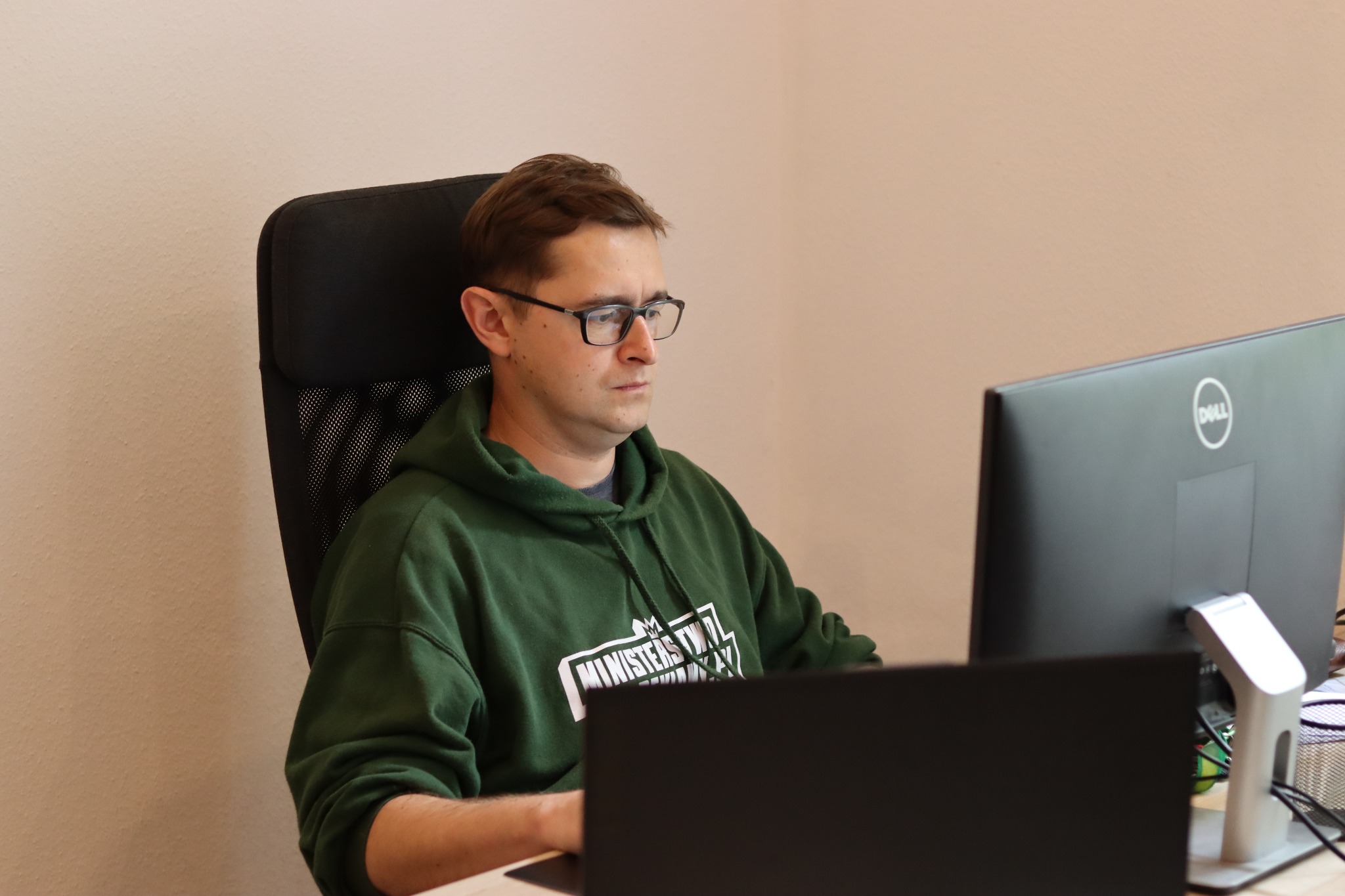 Mężczyzna w okularach i zielonej bluzie skupiony na pracy przy dwóch monitorach, siedząc na czarnym krześle biurowym w jasnym pomieszczeniu.