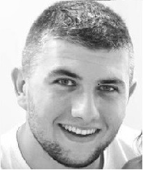 Portret młodego mężczyzny z krótkimi włosami, w czarno-białej tonacji, z delikatnym uśmiechem, skierowany lekko w lewo, na jasnym tle. Widoczne fragmenty ramion.