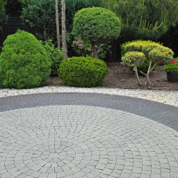 PH.Gregor Grzegorz Pięta - Okrągły, szaro-czarny bruk na tle dekoracyjnych krzewów i drzew w ogrodzie. Kora i białe kamyki uzupełniają aranżację.