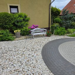 PH.Gregor Grzegorz Pięta - Ogród z białymi kamieniami,  kwitnącymi krzewami i drzewami.  Biała ławeczka i okrągły wzór z kostki brukowej tworzą przytulną i zadbaną przestrzeń.