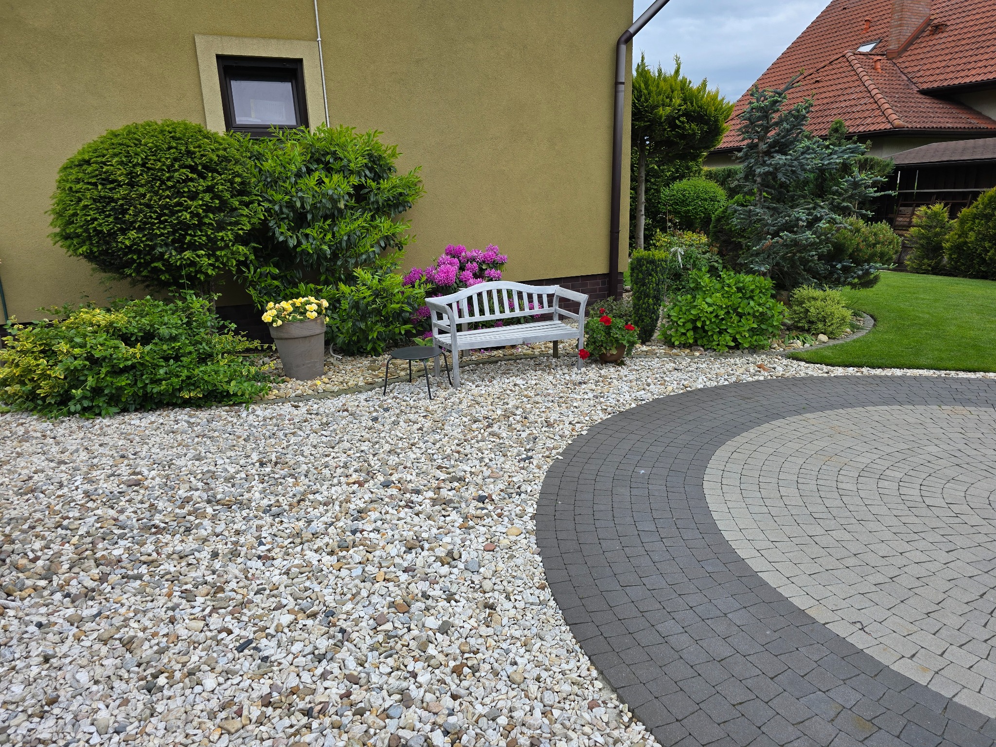 Ogród z białymi kamieniami,  kwitnącymi krzewami i drzewami.  Biała ławeczka i okrągły wzór z kostki brukowej tworzą przytulną i zadbaną przestrzeń.