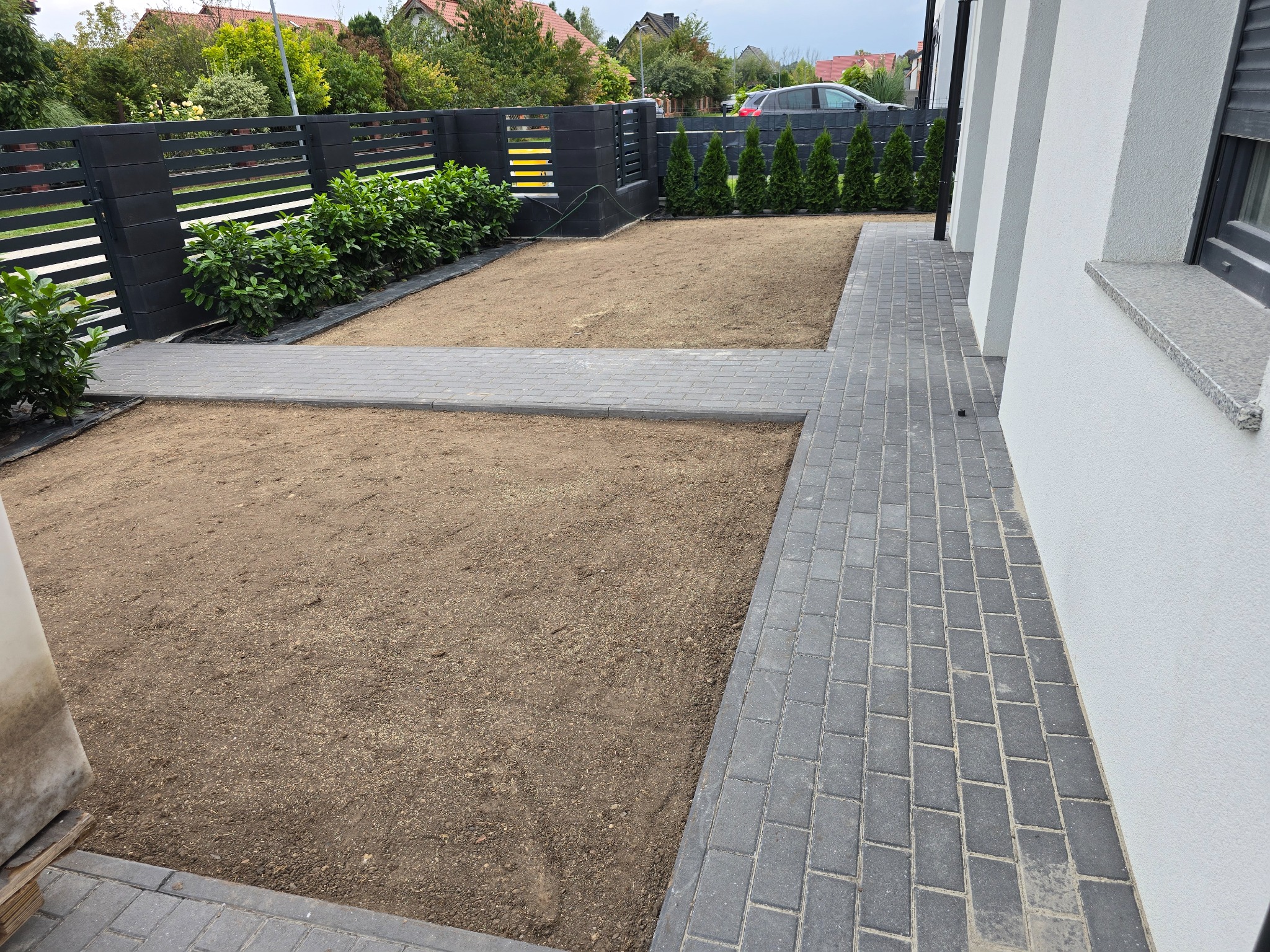 Przygotowany teren pod trawnik z kostką brukową i ozdobnymi krzewami wzdłuż ogrodzenia. Widoczne elementy systemu nawadniania.