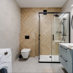 CELMAR Maciej Manelski - Jasna łazienka z kabiną prysznicową w czarnej ramie, umywalką z szafką w kolorze błękitnym i pralką. Na ścianach beżowe płytki ułożone w jodełkę.