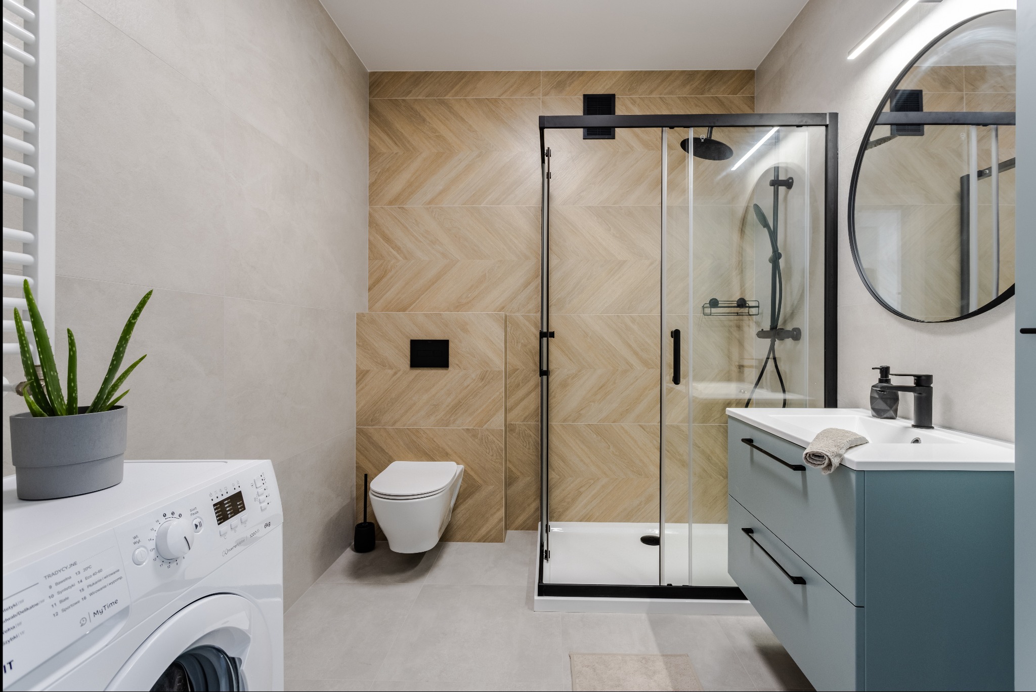 Jasna łazienka z kabiną prysznicową w czarnej ramie, umywalką z szafką w kolorze błękitnym i pralką. Na ścianach beżowe płytki ułożone w jodełkę.