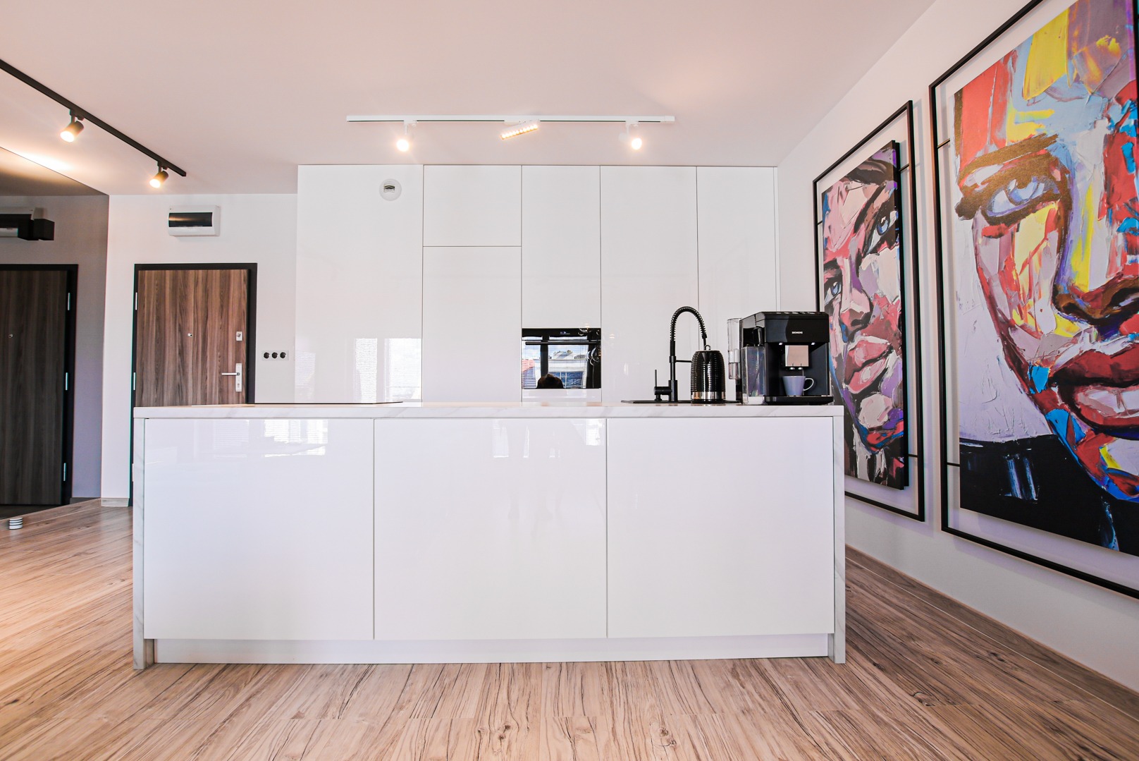 Biała, nowoczesna kuchnia z wyspą, czarną armaturą i ekspres do kawy. Na ścianie abstrakcyjne obrazy. Widoczna drewnopodobna podłoga i drzwi.