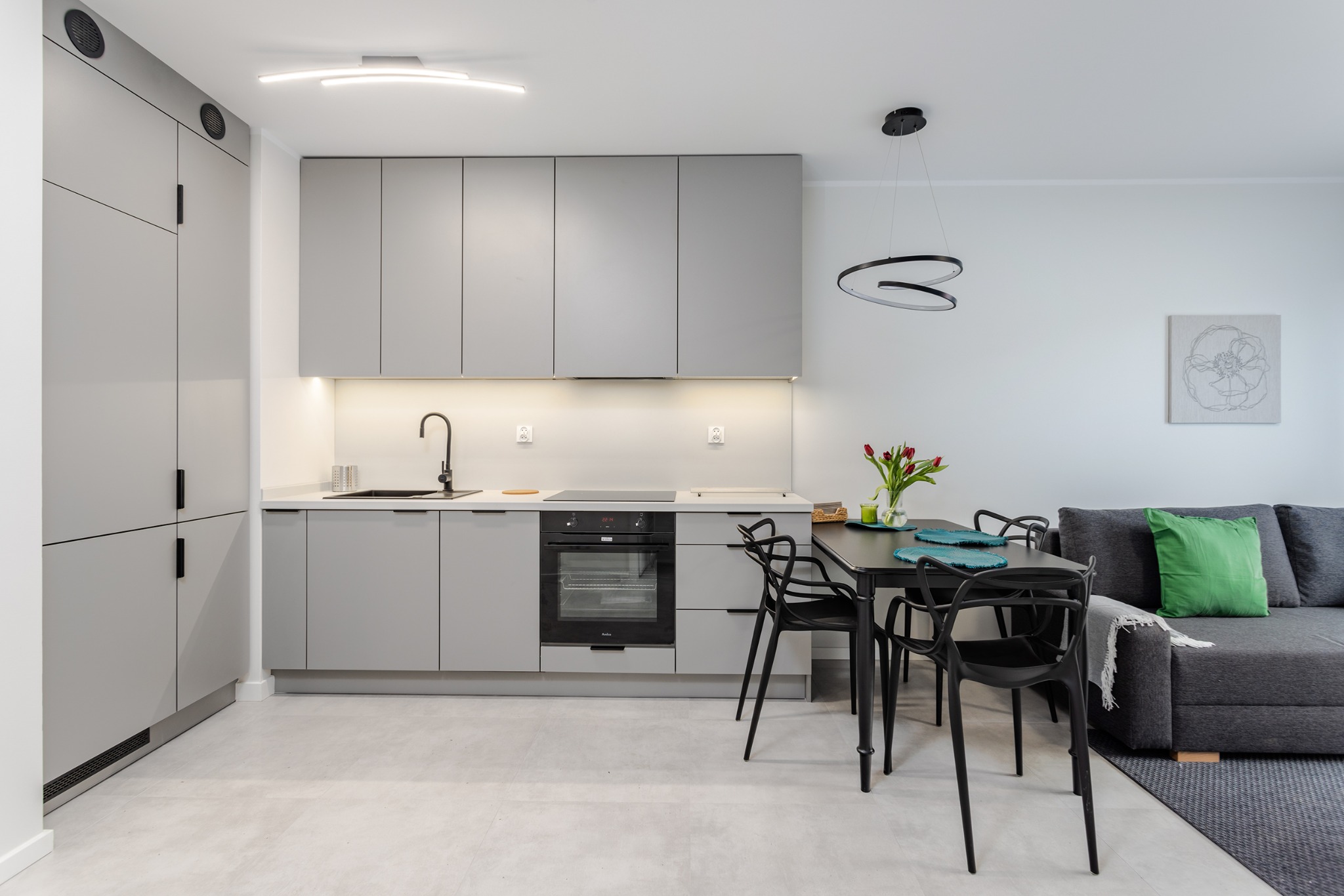 Nowoczesna, szara kuchnia z czarnymi akcentami: bateria, piekarnik, krzesła. Wazon z tulipanami na stole. Minimalistyczny design, jasne wnętrze.