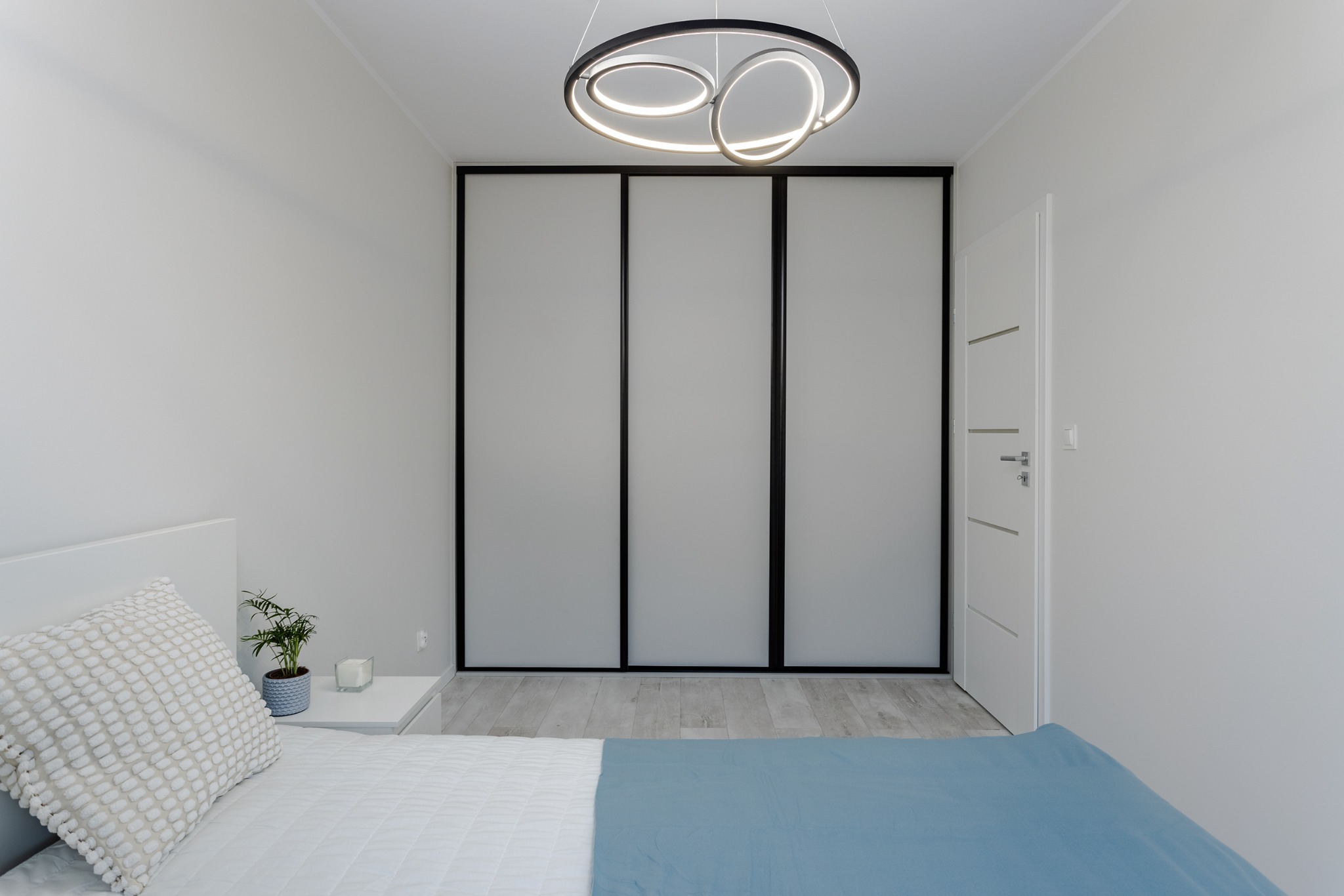 Minimalistyczna sypialnia w bieli z akcentami szarości i błękitu. Widoczne przesuwne drzwi szafy, designerska lampa i fragment łóżka z rośliną na szafce nocnej.