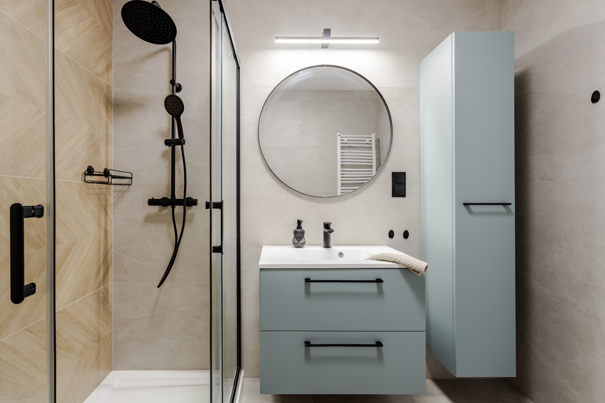 Nowoczesna łazienka z kabiną prysznicową typu walk-in, okrągłym lustrem z podświetleniem i szafką umywalkową w odcieniach błękitu z czarnymi akcentami.