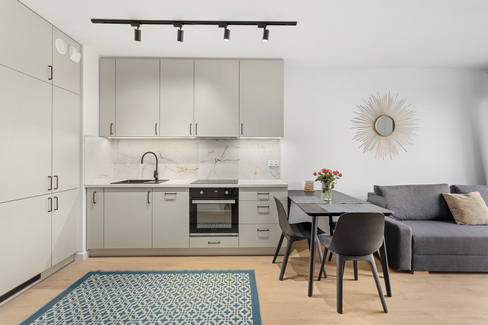 Nowoczesna, jasna kuchnia z szarymi szafkami, marmurowym backsplash'em i drewnianą podłogą. Na stole wazon z różami, obok szara sofa i dekoracyjne lustro.