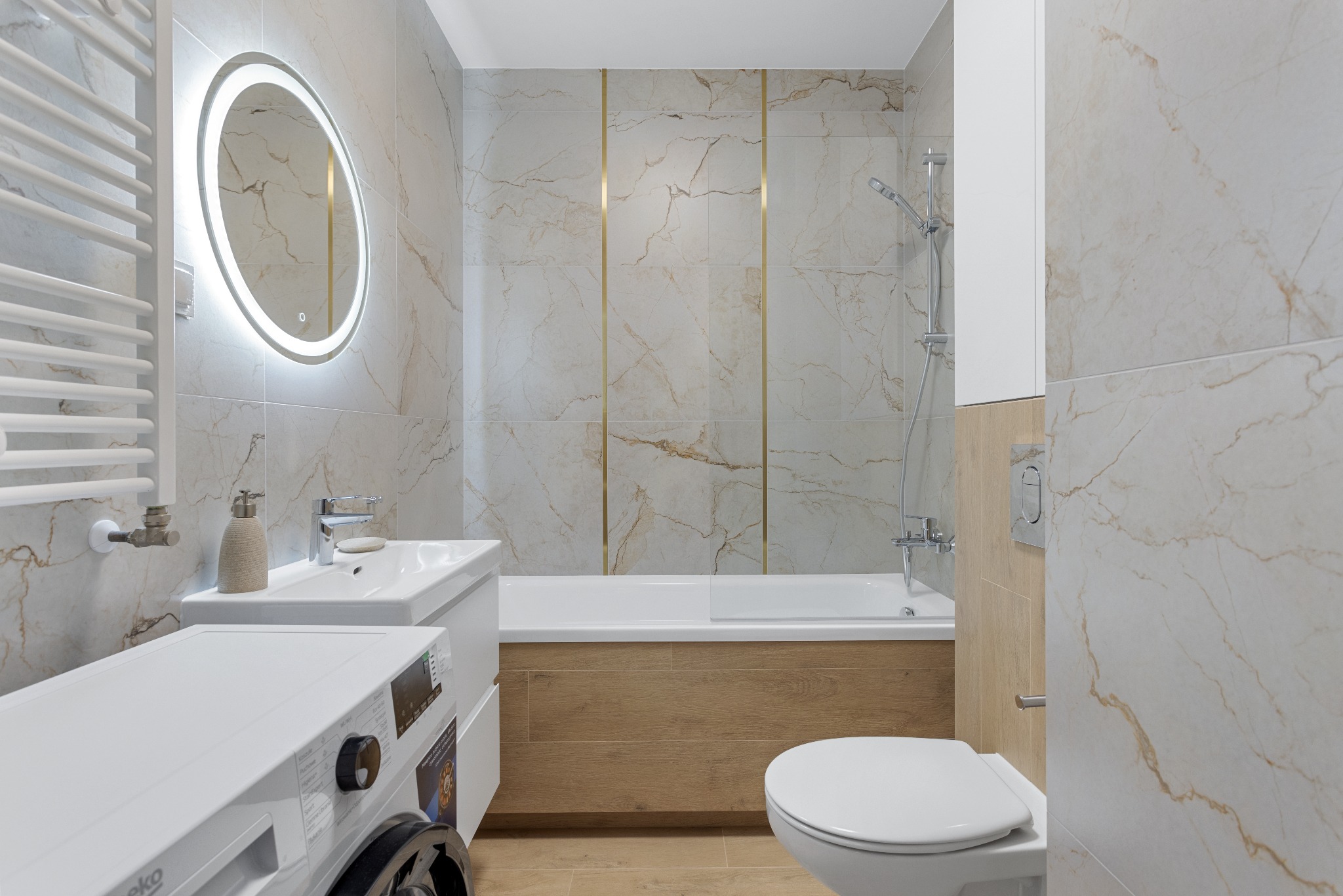Nowoczesna łazienka z marmurowymi płytkami, podświetlanym lustrem i pralką. Uwagę przykuwają złote akcenty między płytkami i drewniane wykończenie wanny.