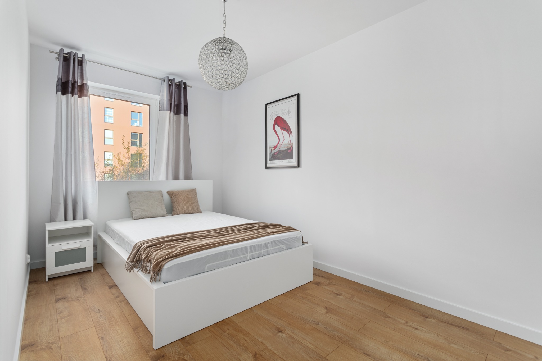 Jasna sypialnia z drewnianą podłogą, białym łóżkiem i szafką nocną. Okno z szarymi zasłonami. Na ścianie plakat z flamingiem i lampa sufitowa.