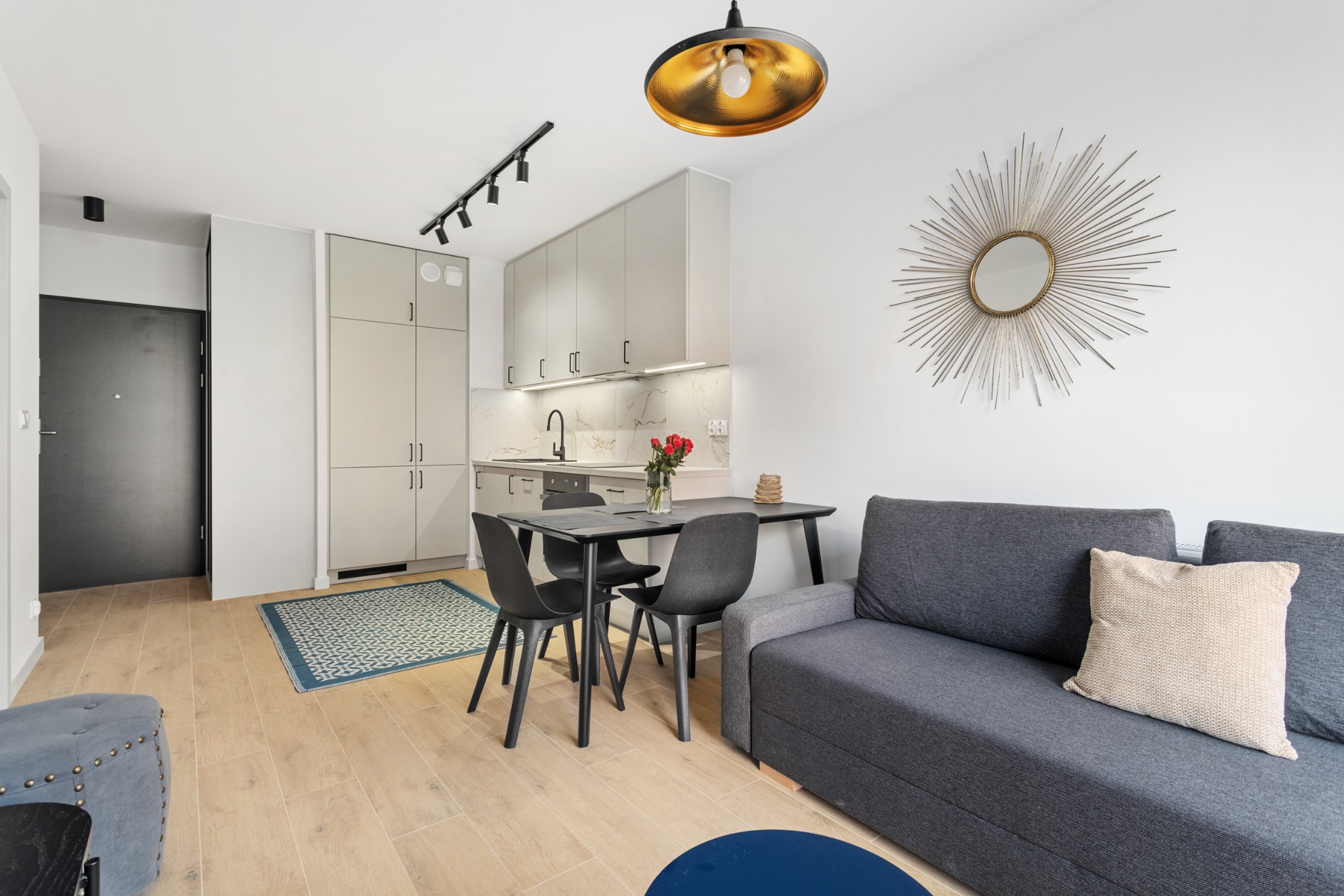 Jasne wnętrze mieszkania: szara sofa, stół z krzesłami, aneks kuchenny z marmurowym blatem i złota lampa. Całość w neutralnej kolorystyce z akcentami złota.