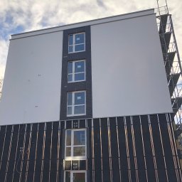 Mykhailo Marusiak - Ocieplanie budynku z widoczną warstwą izolacyjną i rusztowaniem. Fragment elewacji z oknami w trakcie prac wykończeniowych. Widok z dołu na budynek.