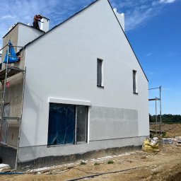 Mykhailo Marusiak - Biała elewacja domu jednorodzinnego w trakcie wykończenia, z widocznymi rusztowaniami i pracownikiem na dachu. Okna zabezpieczone folią, fragment ściany w szarym podkładzie.