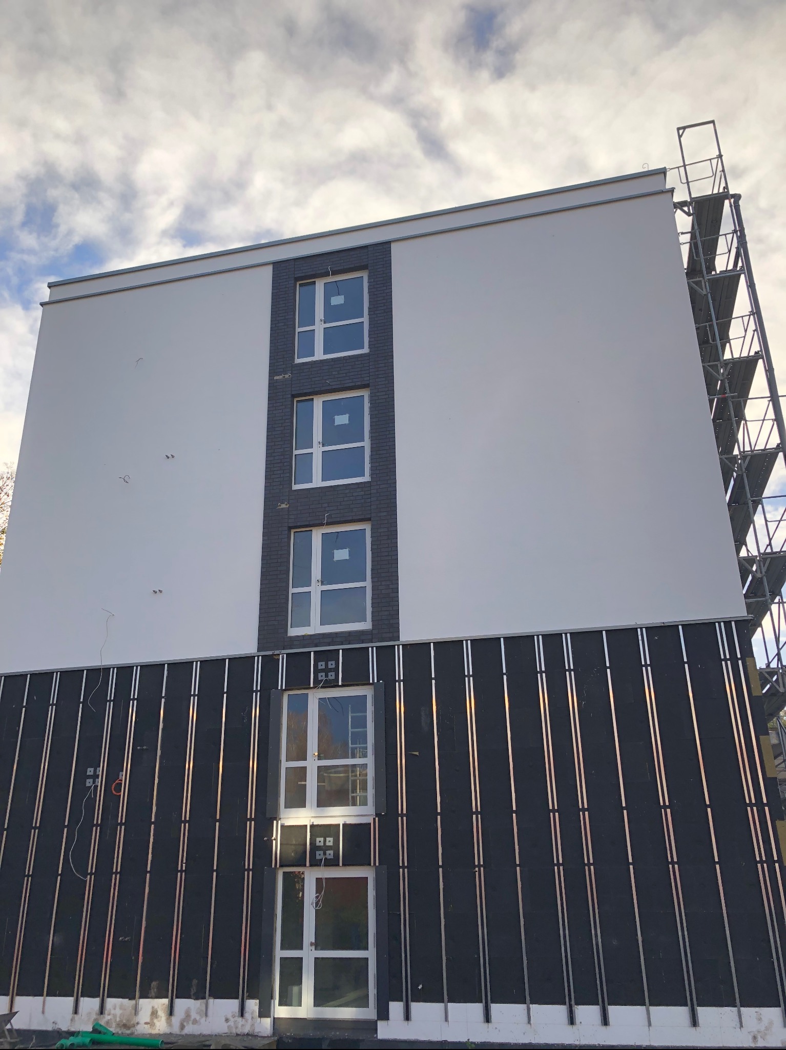 Ocieplanie budynku z widoczną warstwą izolacyjną i rusztowaniem. Fragment elewacji z oknami w trakcie prac wykończeniowych. Widok z dołu na budynek.