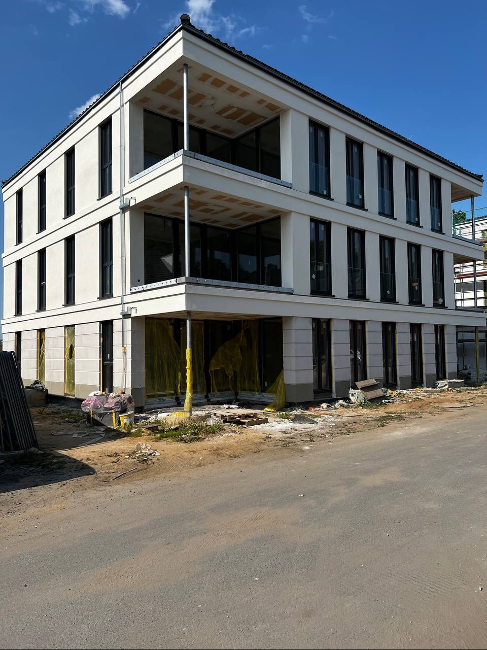 Trzypiętrowy budynek w trakcie wykańczania elewacji, z widocznymi oknami i elementami konstrukcyjnymi. Żółte folie ochronne na parterze. Ujęcie w słoneczny dzień.