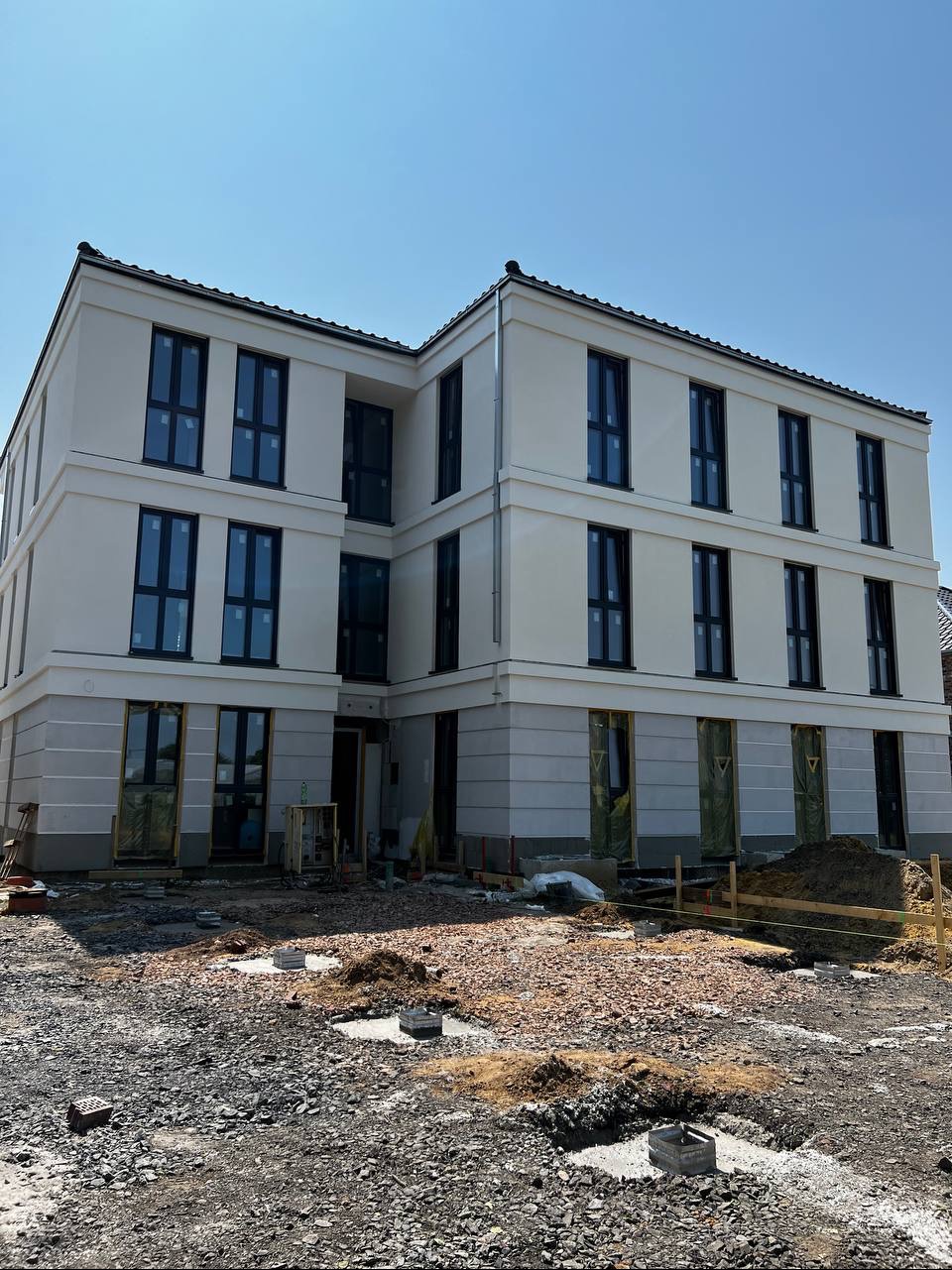 Nowoczesny, trzypiętrowy budynek z jasną elewacją i ciemnymi oknami. Teren wokół budynku w trakcie prac ziemnych, widoczne fundamenty i przygotowania do zagospodarowania.