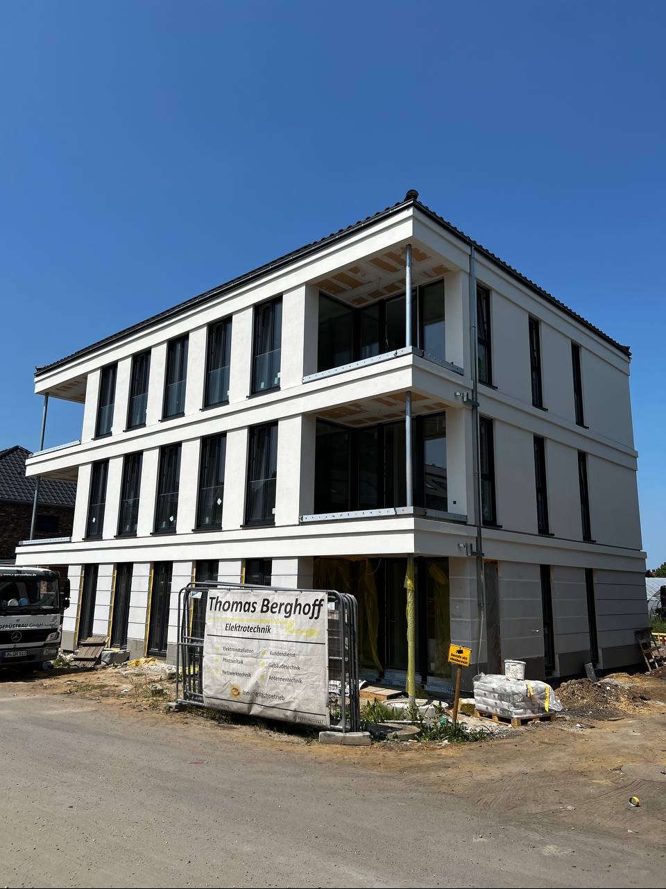 Nowoczesny, trzypiętrowy budynek z białymi elewacjami i czarnymi oknami, w trakcie wykończenia. Widoczny baner firmy 'Thomas Berghoff Elektrotechnik'.