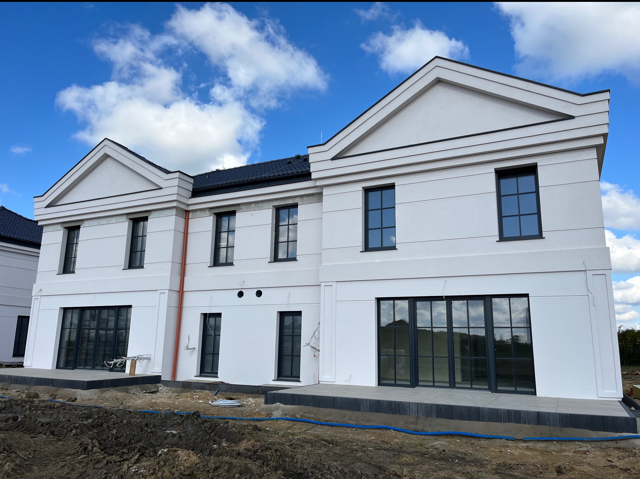 Biała elewacja bliźniaka z czarnymi oknami na tle błękitnego nieba z chmurami. Widoczne elementy wykończeniowe i teren wokół budynku w trakcie prac.