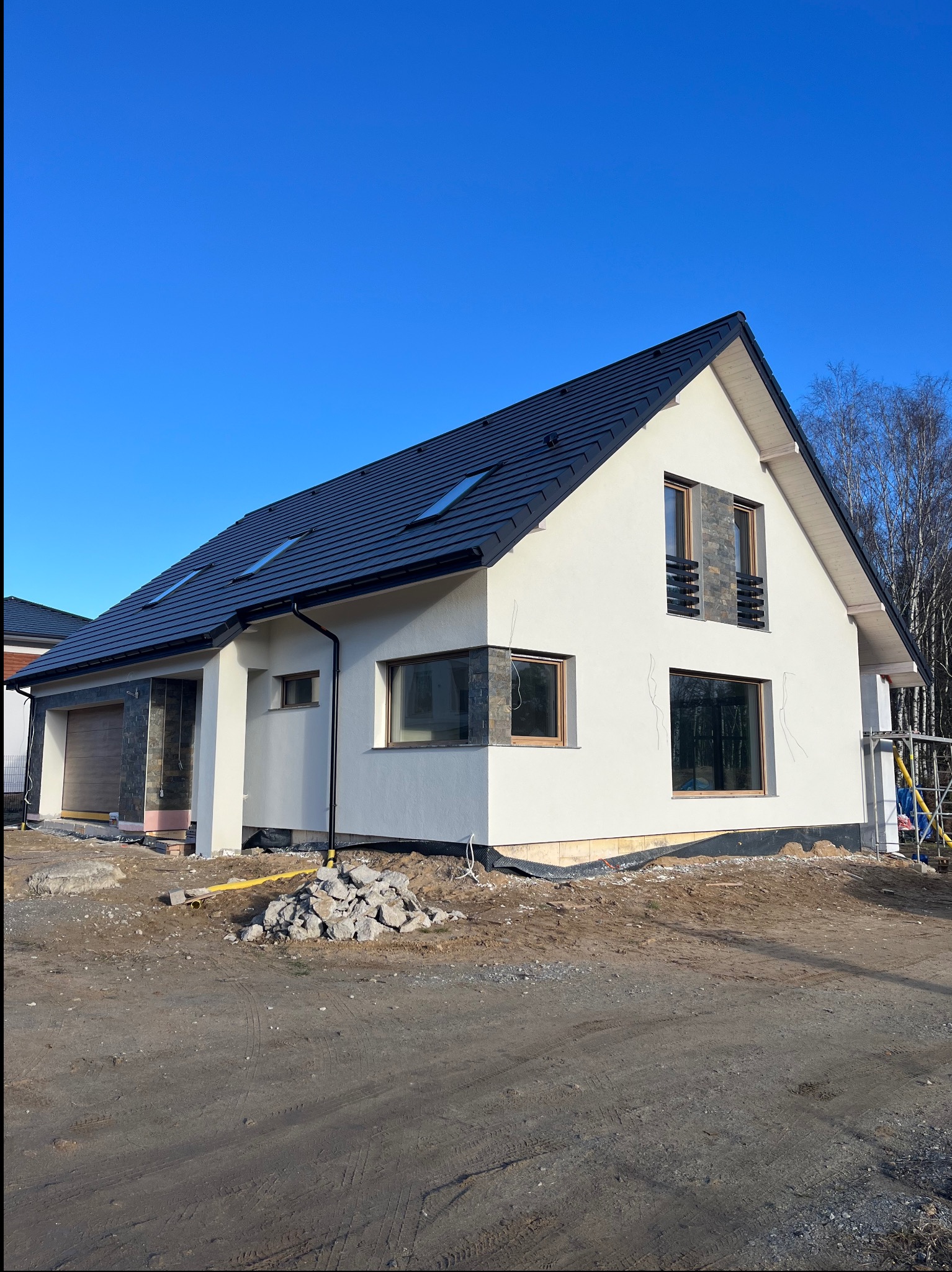 Nowoczesny dom jednorodzinny z białą elewacją i czarnym dachem, wykończony kamieniem dekoracyjnym. Widoczne okna z drewnianą stolarką i fragment rusztowania.