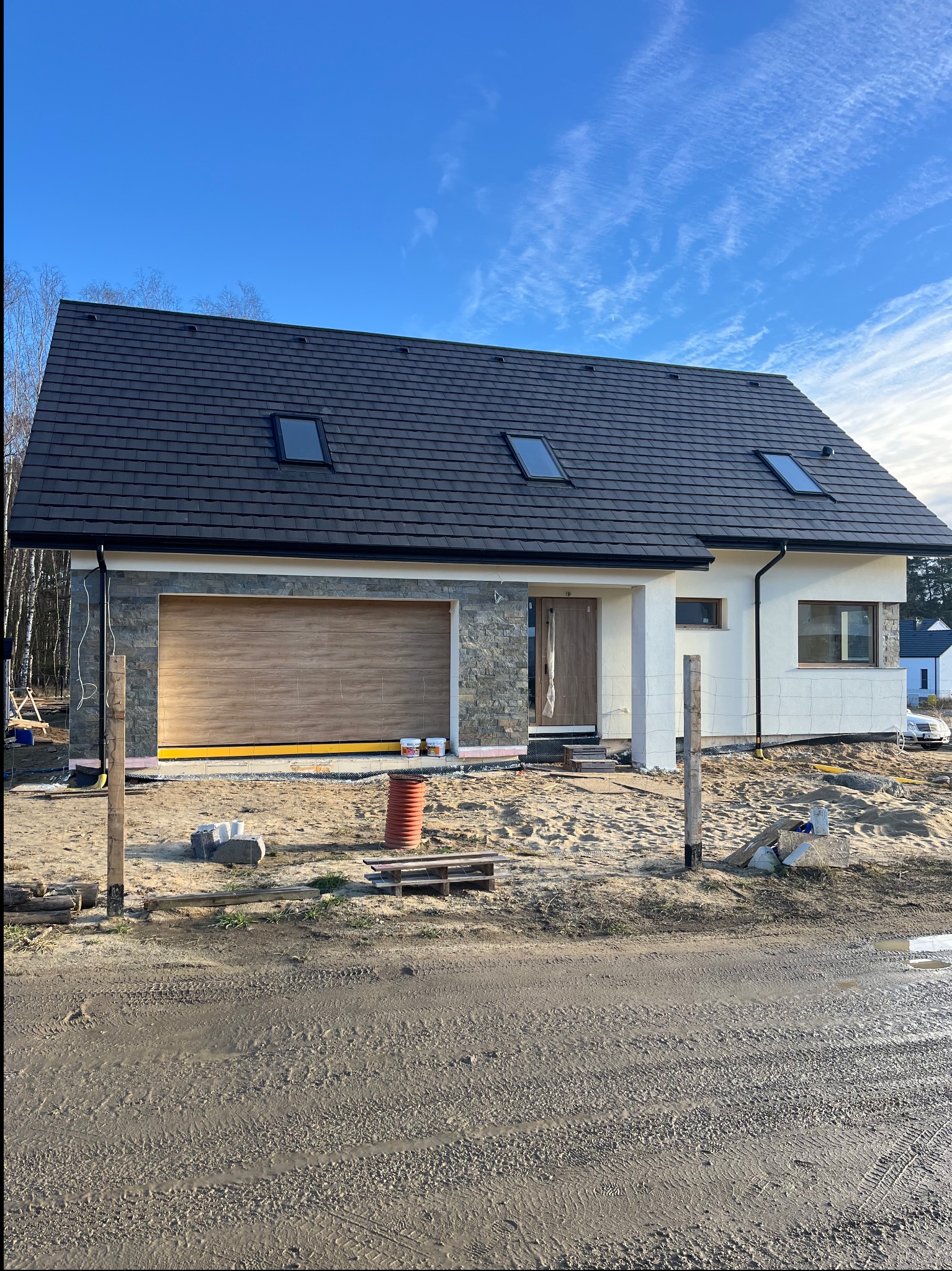 Nowoczesny dom jednorodzinny w trakcie wykończenia elewacji, z widoczną drewnianą bramą garażową i fragmentem ocieplonej ściany. Na pierwszym planie teren budowy.