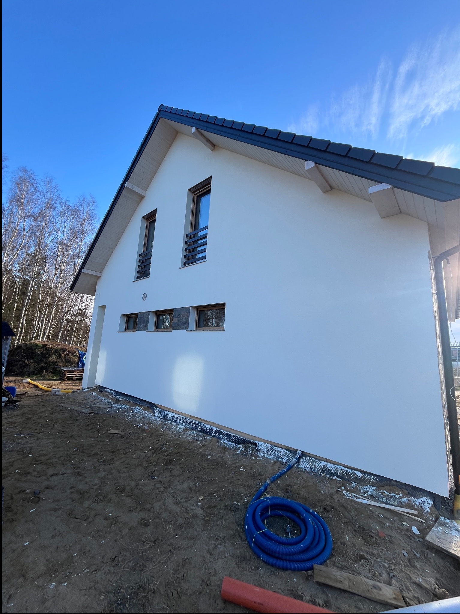 Boczny widok domu z białą elewacją, czarnym dachem i małymi oknami. Wokół domu widoczny teren budowy z niebieskim wężem. Błękitne niebo w tle.