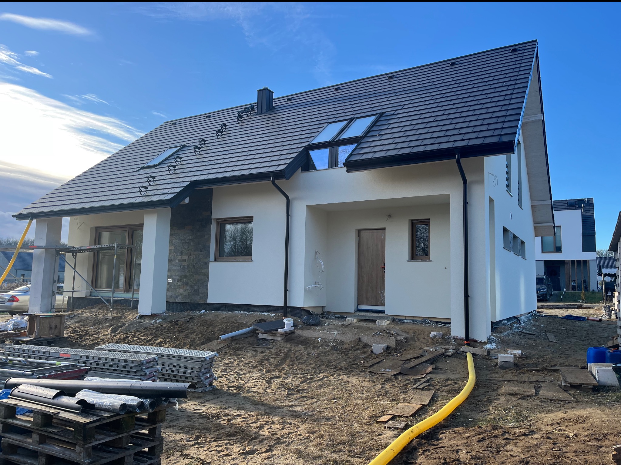 Nowoczesny dom jednorodzinny z białymi ścianami i ciemną dachówką, widoczne okna dachowe. Częściowo wykończona elewacja, teren wokół w trakcie prac ziemnych.