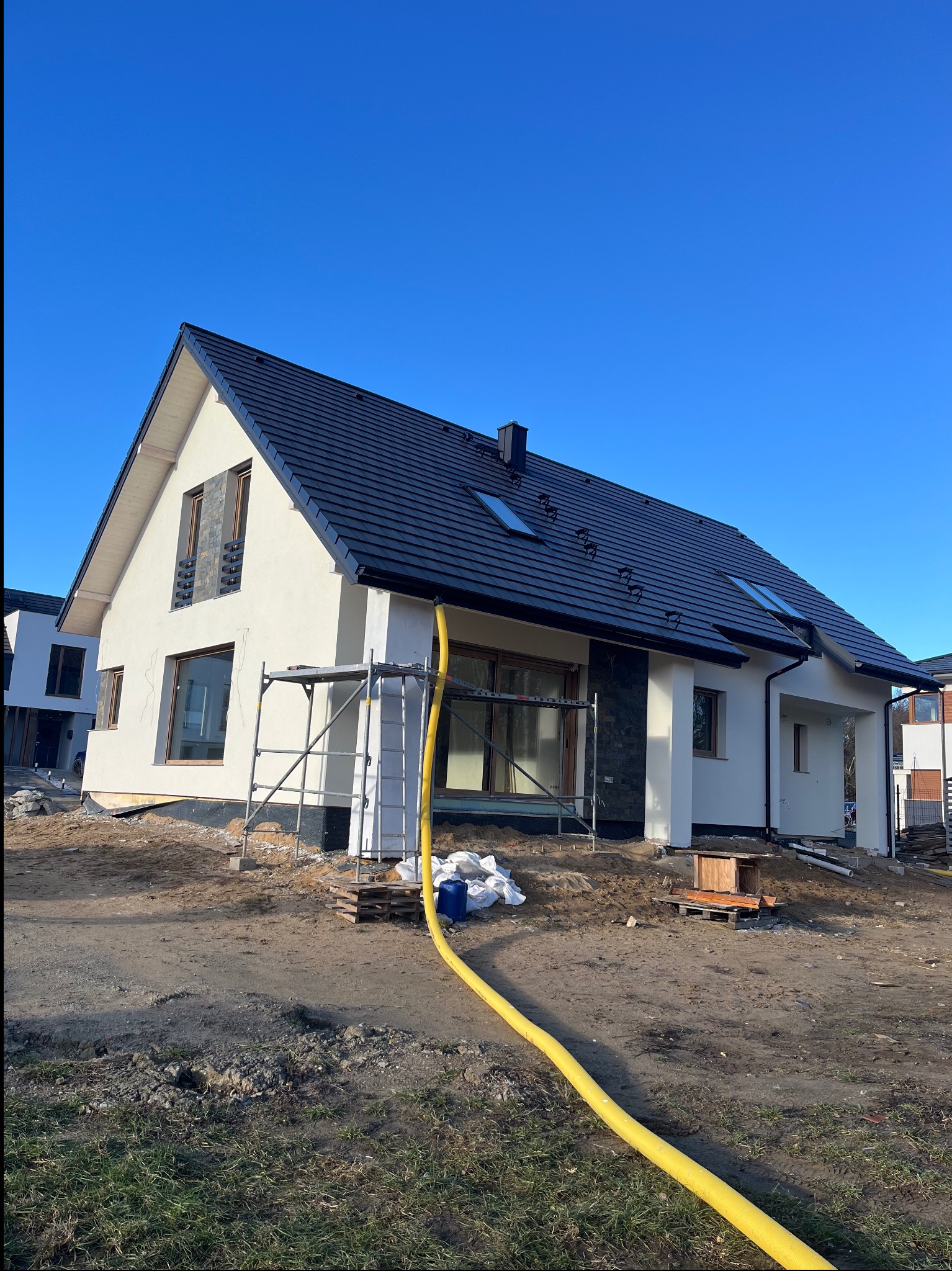 Nowoczesny dom jednorodzinny z czarnym dachem i białą elewacją w trakcie wykończenia. Widoczne rusztowania i żółty wąż. Błękitne niebo w tle.