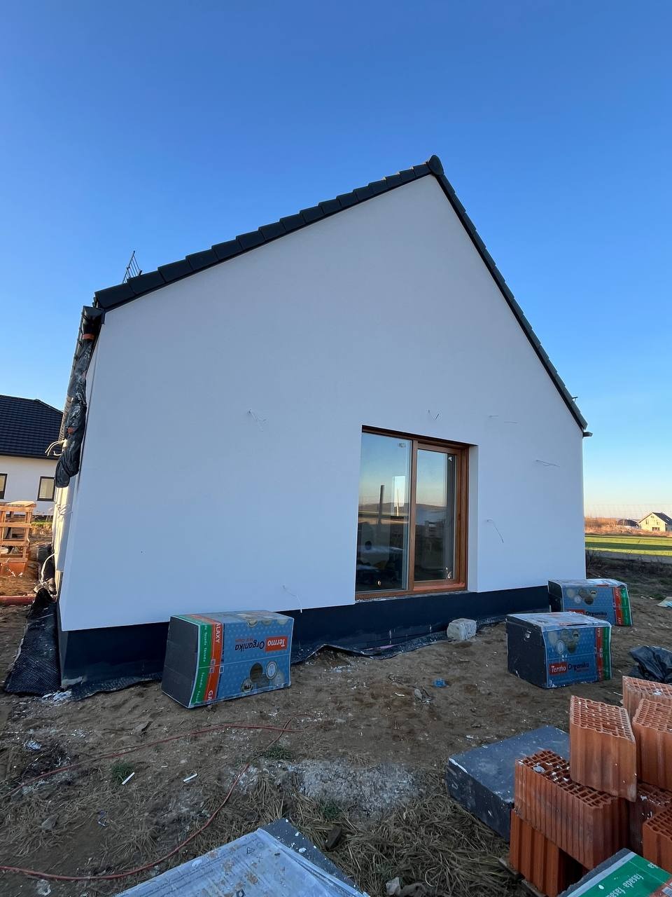 Boczny widok domu z oknem i świeżo otynkowaną, białą elewacją. Na ziemi leżą materiały budowlane, w tym cegły i opakowania styropianu.