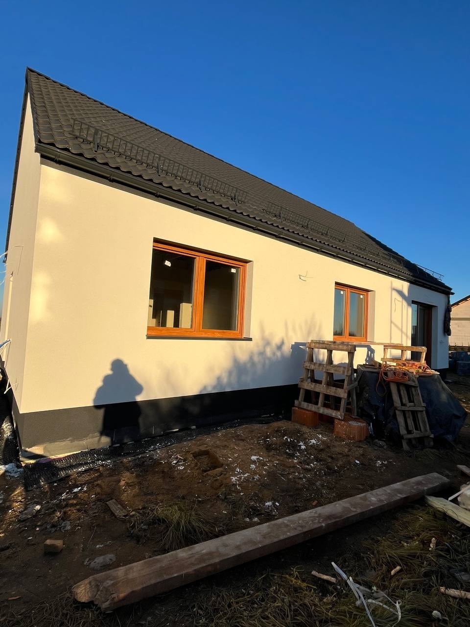 Nowoczesny dom jednorodzinny z jasną elewacją i ciemnym dachem, widoczne drewniane okna, teren wokół domu w trakcie prac wykończeniowych, cień osoby na ścianie.