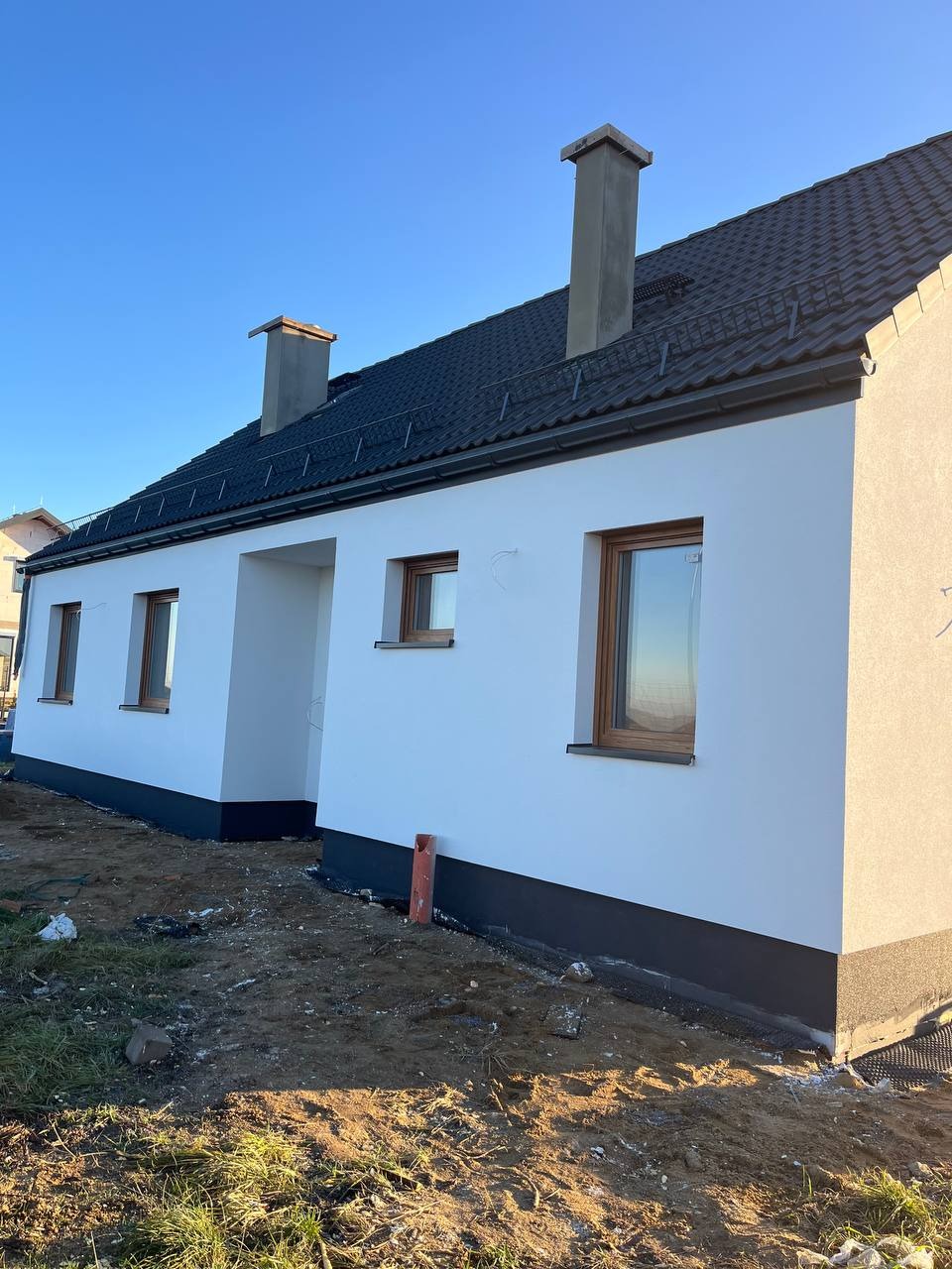 Boczny widok domu z białą elewacją, brązowymi oknami i ciemnym dachem. Widoczny fragment terenu przed budynkiem i błękitne niebo. Elewacja świeżo pomalowana.