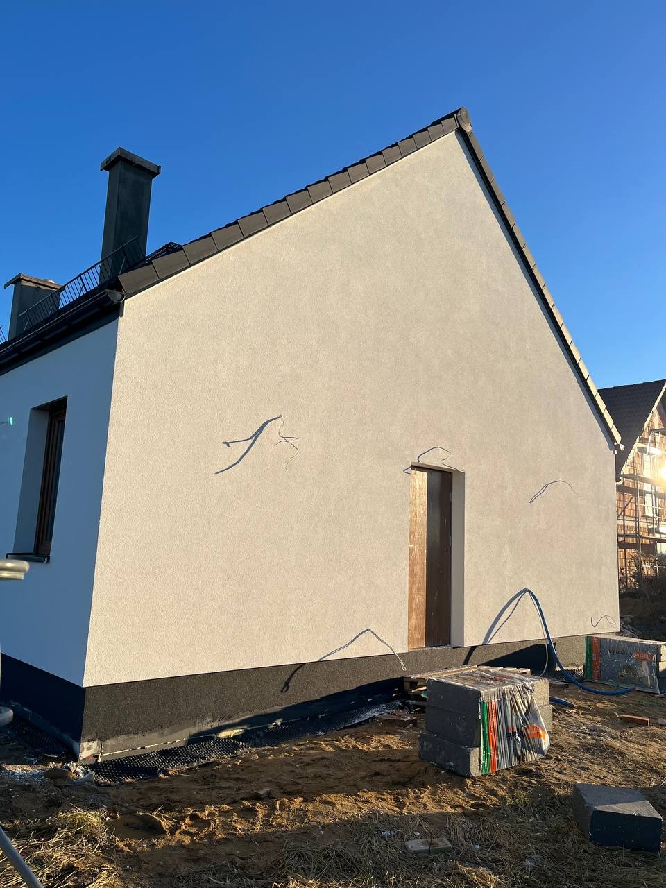 Boczny widok otynkowanego domu jednorodzinnego z czarną podmurówką i dachem. Na ziemi leżą bloczki i zwinięty wąż. Słoneczny dzień.