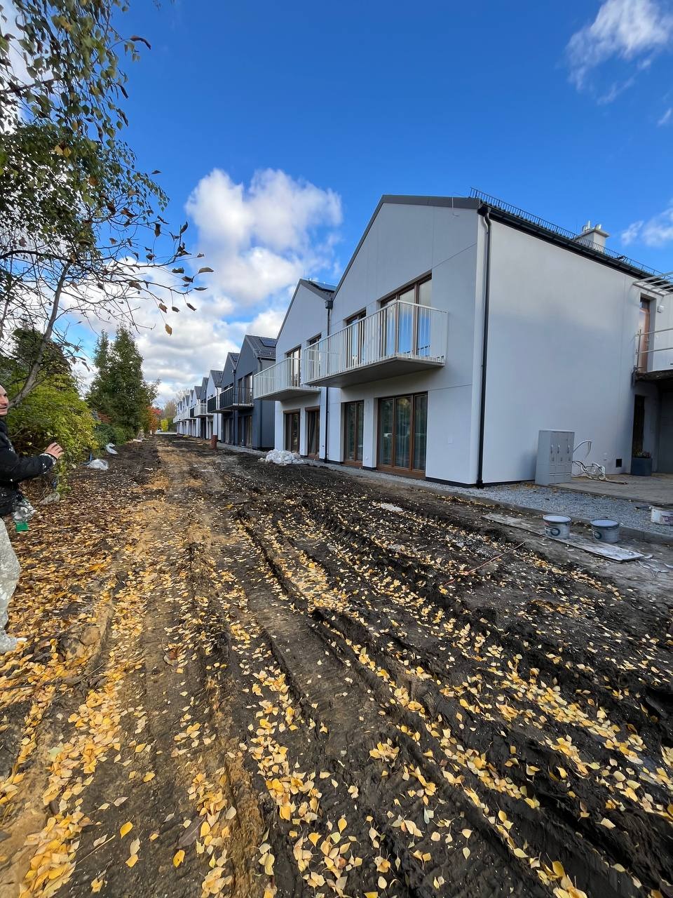 Rząd nowoczesnych domów jednorodzinnych z balkonami, świeżo po wykonaniu elewacji, z widocznymi śladami prac budowlanych na błotnistej drodze.