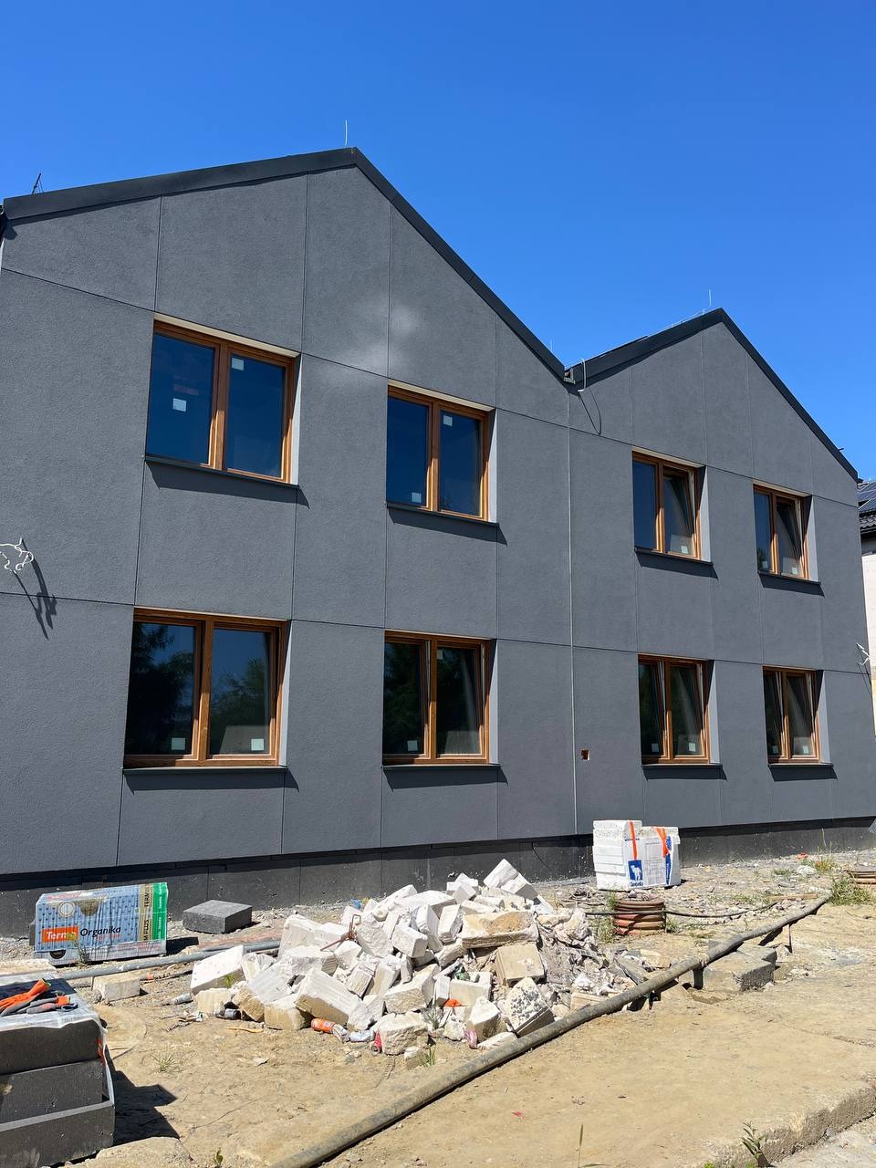 Dwukondygnacyjny budynek z szarą elewacją panelową i drewnianymi oknami. Na pierwszym planie gruz i materiały budowlane.