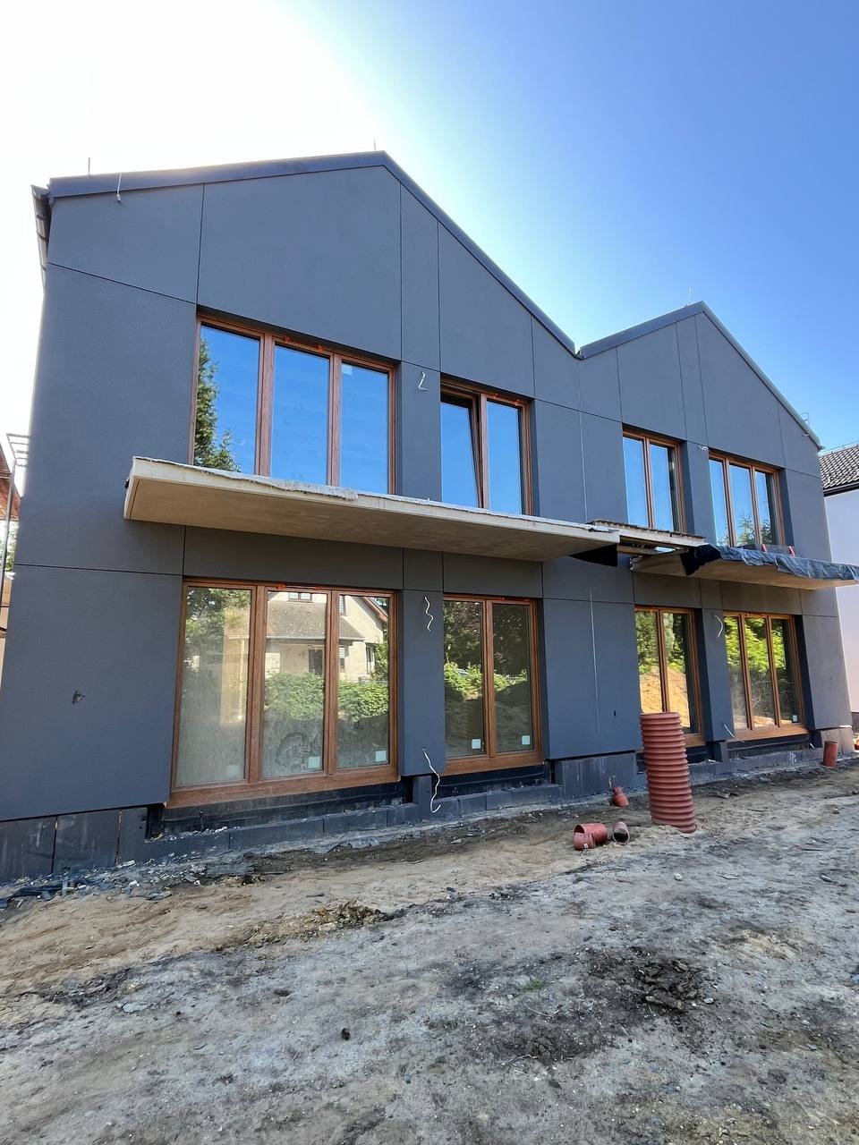 Nowoczesny dom z szarą elewacją panelową i drewnianymi oknami. Widoczne rury kanalizacyjne przy fundamencie. Częściowo wykończona elewacja, surowy teren.
