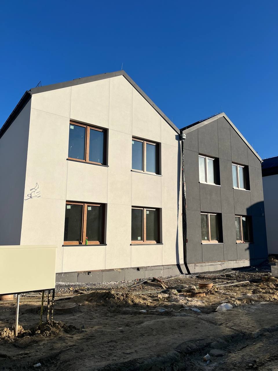 Dwa budynki w trakcie wykańczania elewacji: jeden jasny z drewnianymi oknami, drugi ciemny z białymi. Widoczne niedokończone otoczenie i błękitne niebo.